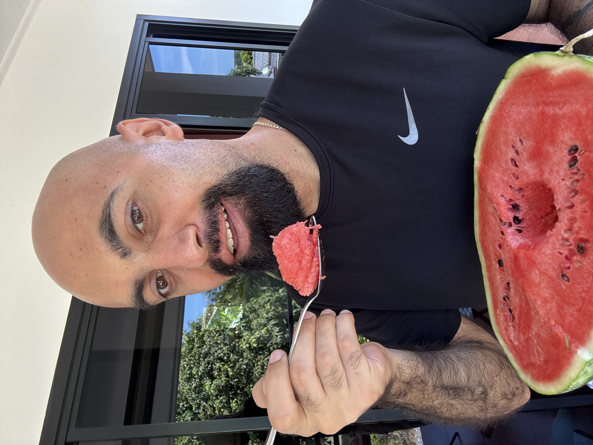 A bald, dark-haired man with a beard is holding a spoonful of watermelon in front of his mouth. He is wearing a black t-shirt with a Nike logo. In front of him is a halved watermelon. The scene appears to be outdoors, possibly on a balcony or patio, with trees and a building visible in the background. The sky is blue. The man is smiling and appears to be enjoying the watermelon. The location is the city of Ko Samui District, Thailand.