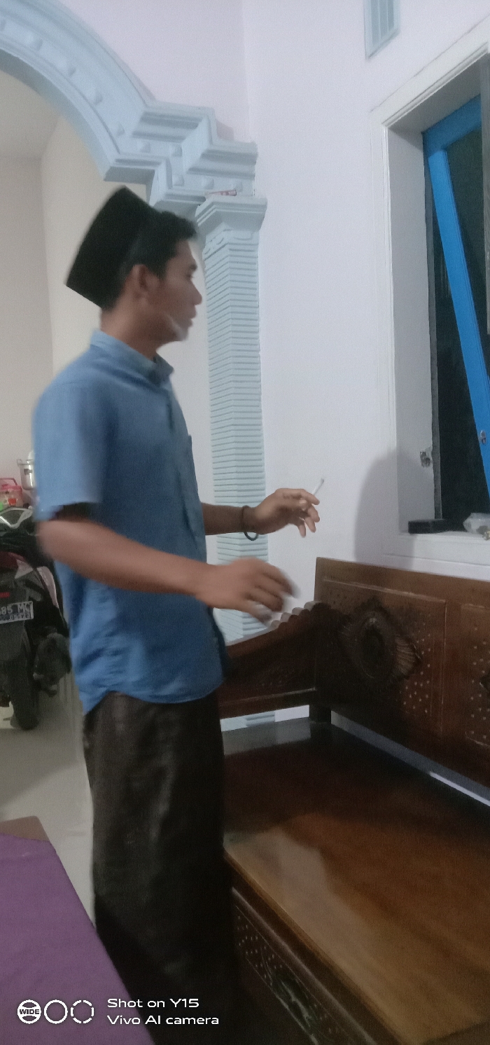 The image displays an indoor scene in Brumbungan Kidul, Indonesia. A male individual, appearing adult, stands in profile, facing right. He wears a light blue short-sleeved collared shirt, dark trousers or sarong, and a black peci on his head. His right hand holds a lit cigarette between his index and middle fingers; his left hand is slightly raised with fingers splayed.
The background on the left features a white wall with an arched doorway adorned with light blue decorative molding and a fluted pillar. To the right of the individual, a white-framed window with a partially visible blue frame is present. A dark brown wooden bench or low table, featuring carved details on its side, extends across the lower right foreground. A purple textile, possibly a mat, is visible on the floor in the lower-left corner. In the background, behind the individual and to the left, a portion of a motorbike, a silver container, and other indistinct objects are discernible. The environment appears to be artificially lit.