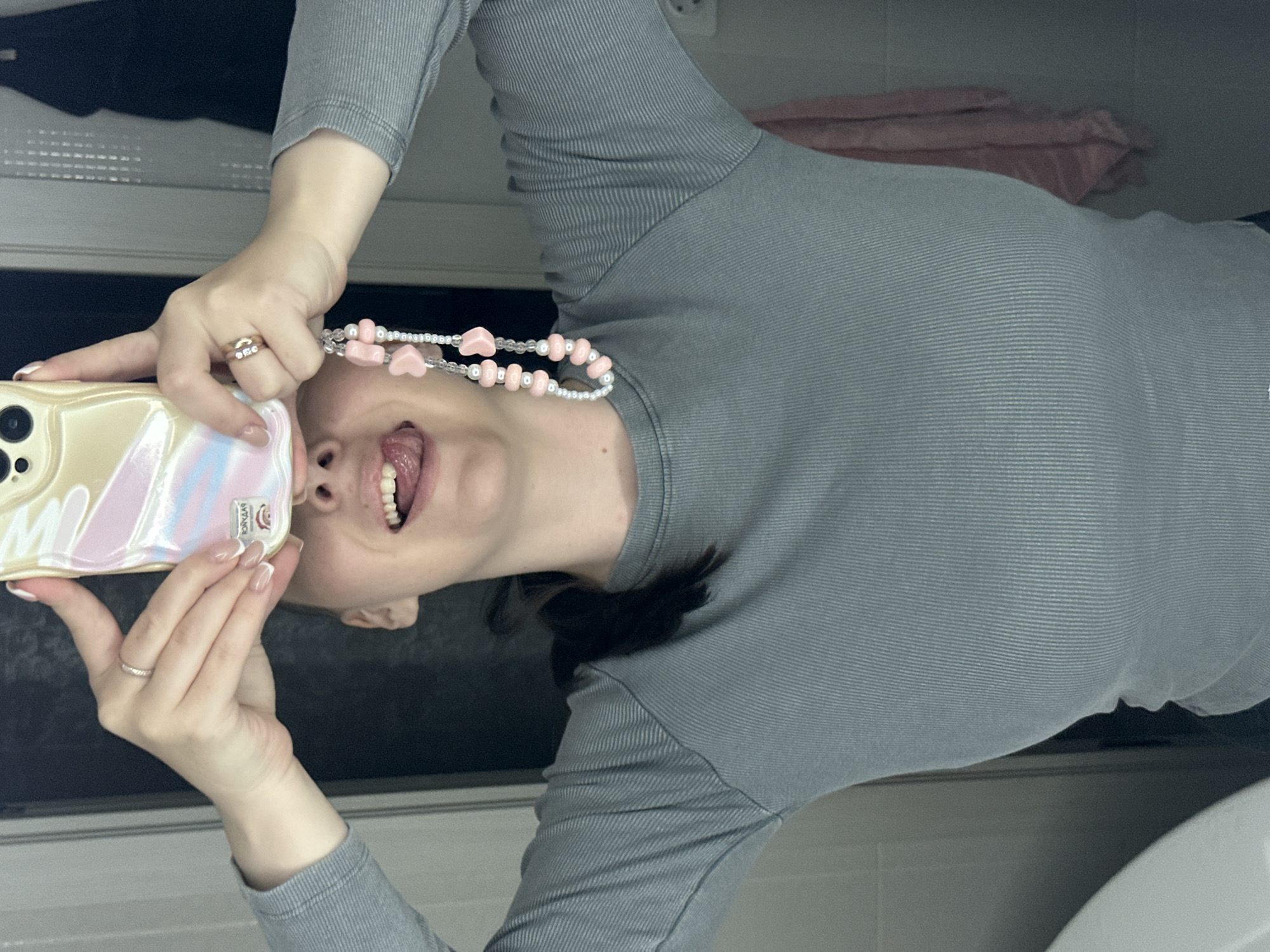 A woman is indoors, holding a cell phone, and protruding her tongue. She is wearing a gray shirt with a pink beaded necklace. The location cannot be determined as Chaikovsky, Russia based on the image.