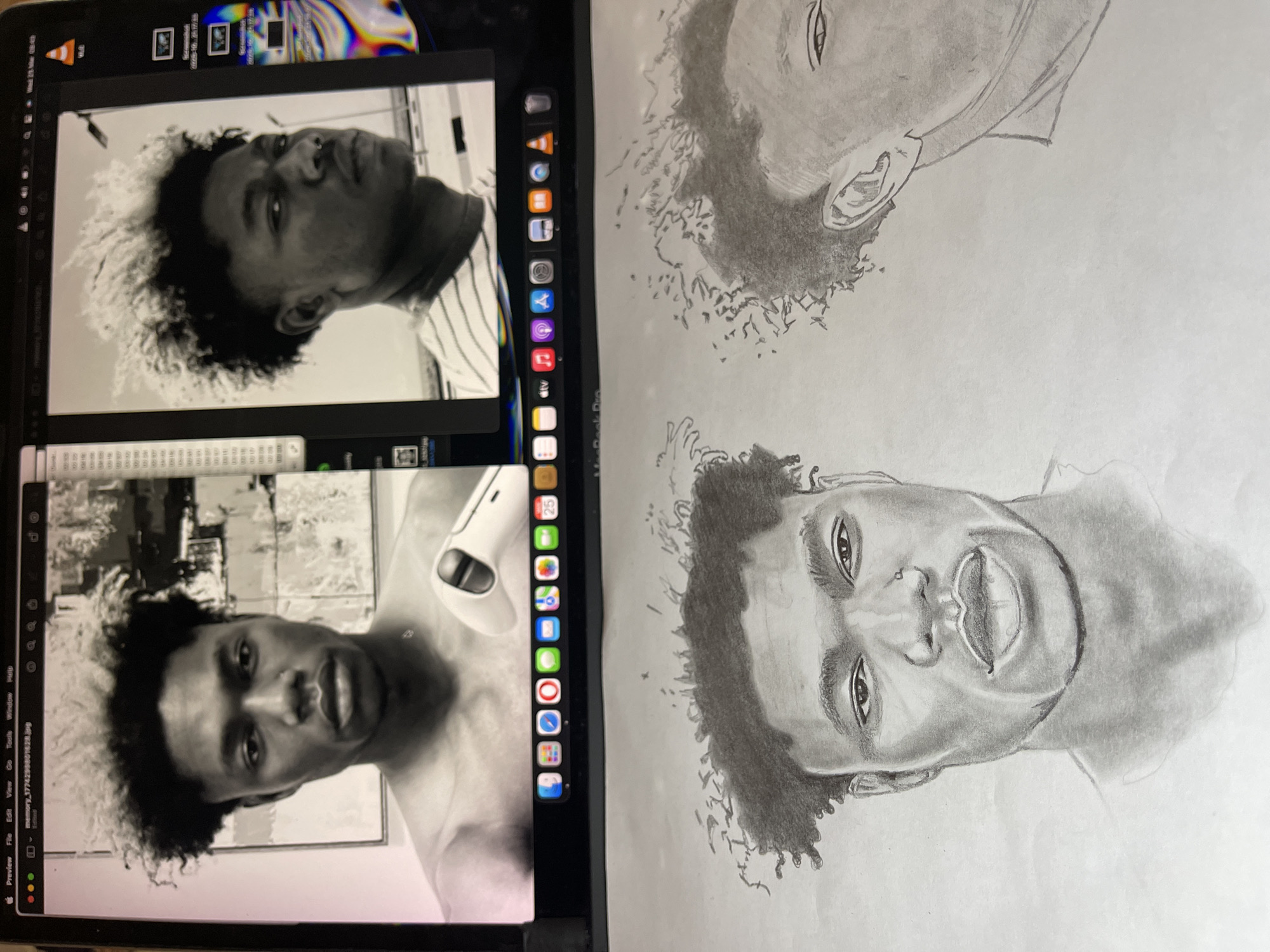 The image depicts an indoor scene in the city of Jos, Nigeria, featuring a MacBook Pro laptop positioned above a sheet of paper containing pencil sketches. The laptop screen displays two black-and-white digital portraits of a young male, which serve as references for the artwork below.
The left portion of the laptop screen shows a front-facing, head-and-shoulders portrait of a male subject with curly, textured hair that appears lighter at the tips, and bare shoulders. A partial white game controller is visible in the lower right background of this digital image. The right portion of the screen displays a profile view (facing right) of the same male, wearing a striped shirt and featuring a visible nose ring, set against a lighter background. The macOS desktop environment, including the "Preview" application interface and an application dock with various icons, is visible across the bottom of the laptop screen.
On the paper below the laptop, two detailed monochrome pencil sketches of the male subject are present. The left sketch is a full front-facing portrait, closely replicating the features and expression from the corresponding digital image above. To its right, a partial sketch of the male's head and upper chest in profile is visible, corresponding to the right digital portrait. The scene illustrates an artistic process involving the transfer of digital photographic references into hand-drawn artwork.