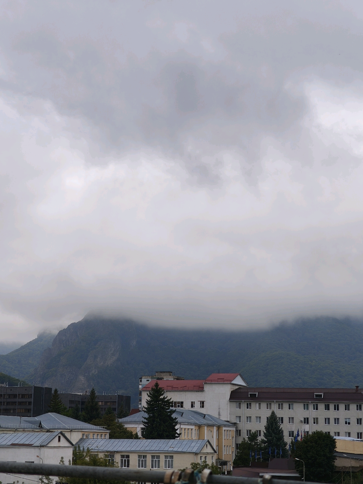 The image shows a view of Karachayevsk, Russia, with buildings in the foreground and a mountain range in the background. The sky is overcast with heavy clouds obscuring the upper portions of the mountains. The buildings appear to be a mix of residential and institutional structures, with multiple stories and varying architectural styles. Some buildings have red roofs, while others have gray or brown roofing. Trees are interspersed among the buildings, with evergreen trees being prominent. There are no people visible in the image. The overall impression is a somewhat gloomy or overcast day in a mountainous city.