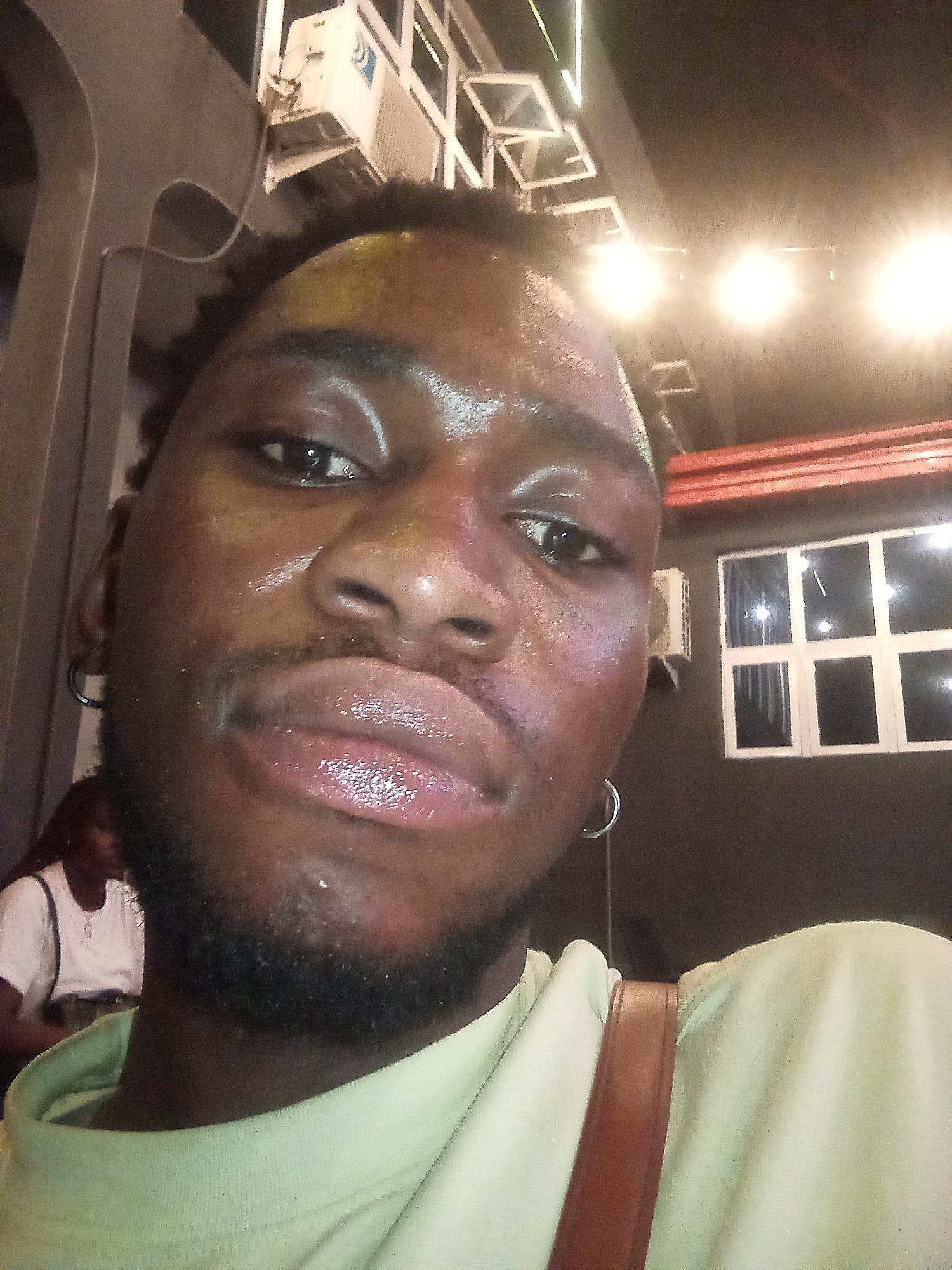 A selfie of a Black man with short hair and a beard, wearing a light-colored shirt and a brown strap across his shoulder. He is wearing silver hoop earrings. His face appears to be sweaty or oily. Behind him are overhead lights, an air conditioning unit, and a window with multiple panes. To the left, there is a woman with dark hair wearing a white shirt. The location is indoors, likely a commercial establishment. There is no verifiable information that states the location of this image in Lokoja, Nigeria.