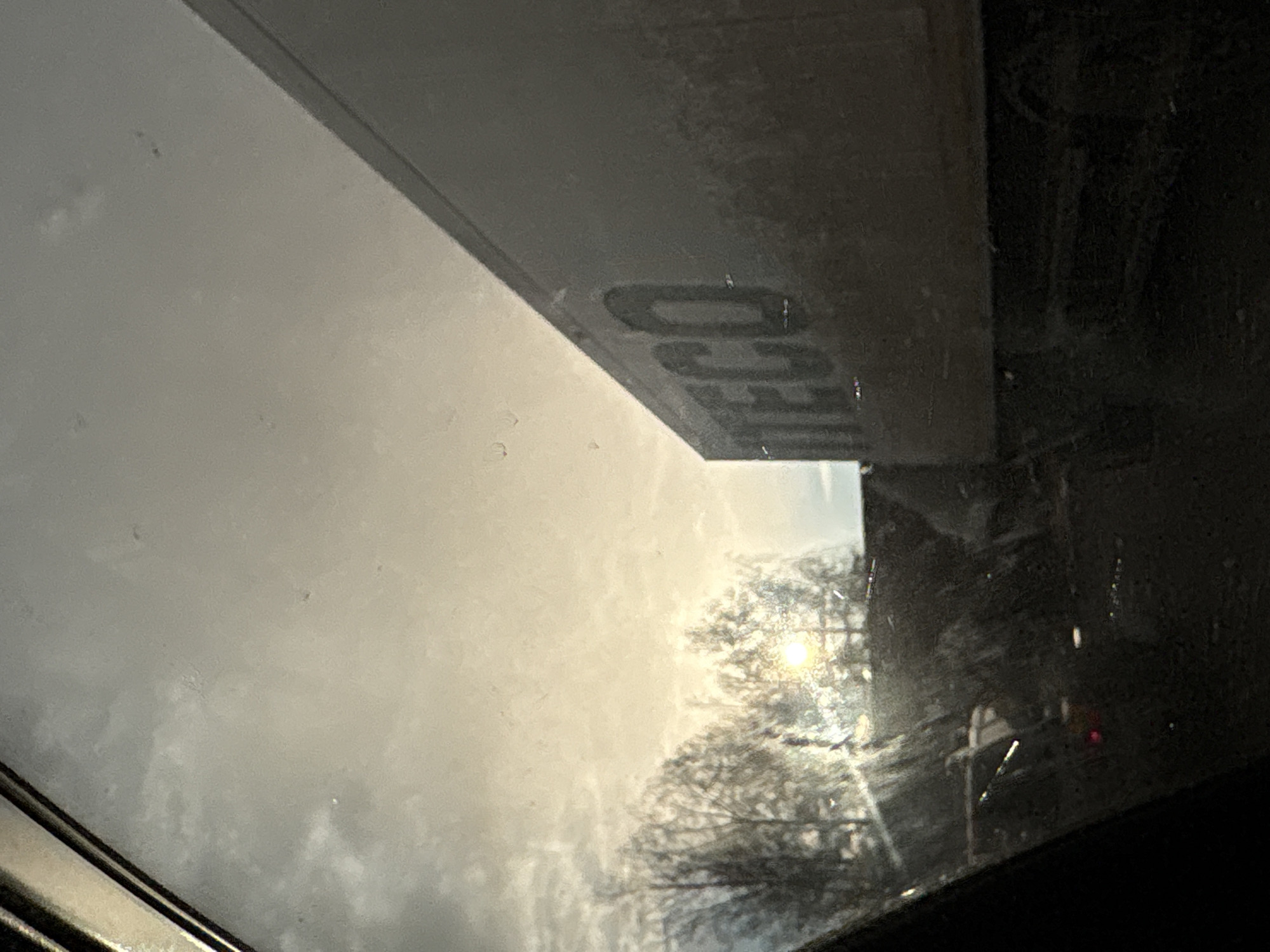 The image depicts an outdoor scene captured from inside a vehicle, looking through a window that exhibits visible dust and streaks. The perspective is tilted, but when viewed upright, a large, light-colored rectangular canopy or overhang structure is prominent in the upper part of the frame. On its underside, dark, raised lettering is visible, clearly spelling "DECO".
The mid-left section of the image shows an overcast sky with light grey clouds. In the mid-right, a bright, low-lying sun shines through the silhouettes of numerous bare tree branches, creating a strong glare and suggesting either early morning or late afternoon. The light casts a warm, soft glow on the cloudy sky. The lower and far-right areas are darker, possibly showing parts of the car's interior or obscured ground details. No people are visible. The scene evokes a moment of observation from a vehicle, possibly at a gas station or a similar drive-through location in Kazan, Russia.