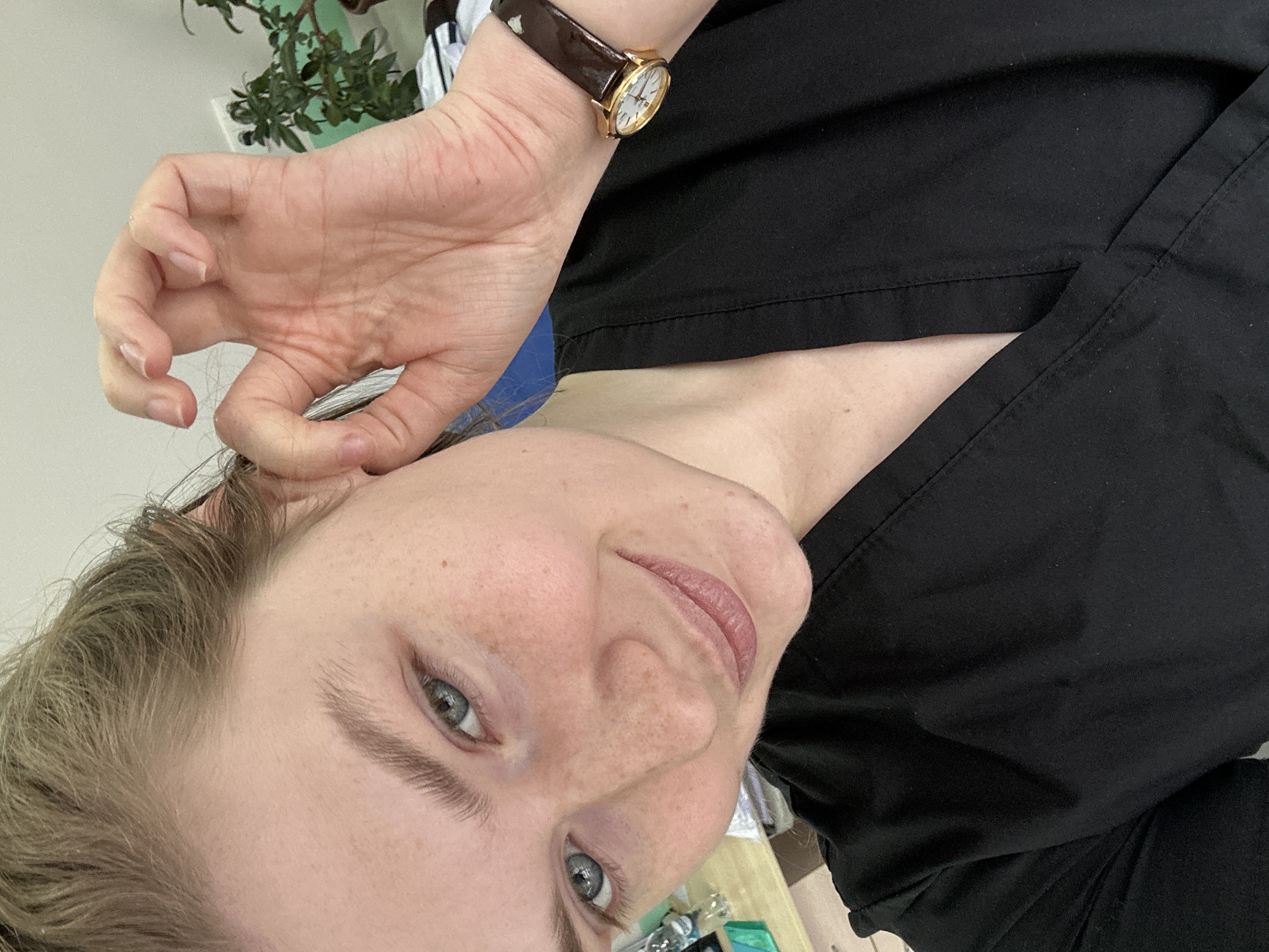 A close-up, selfie-style photograph shows a person with fair skin, freckles, light brown hair, and blue eyes. They are wearing a black collared shirt. The person's left hand is raised near their left ear, with their thumb and index finger forming a circle. A wristwatch with a brown leather band and a gold face is on their left wrist. The background is blurred, but a green leafy plant is visible in the upper left corner. The location is not discernible from the image.