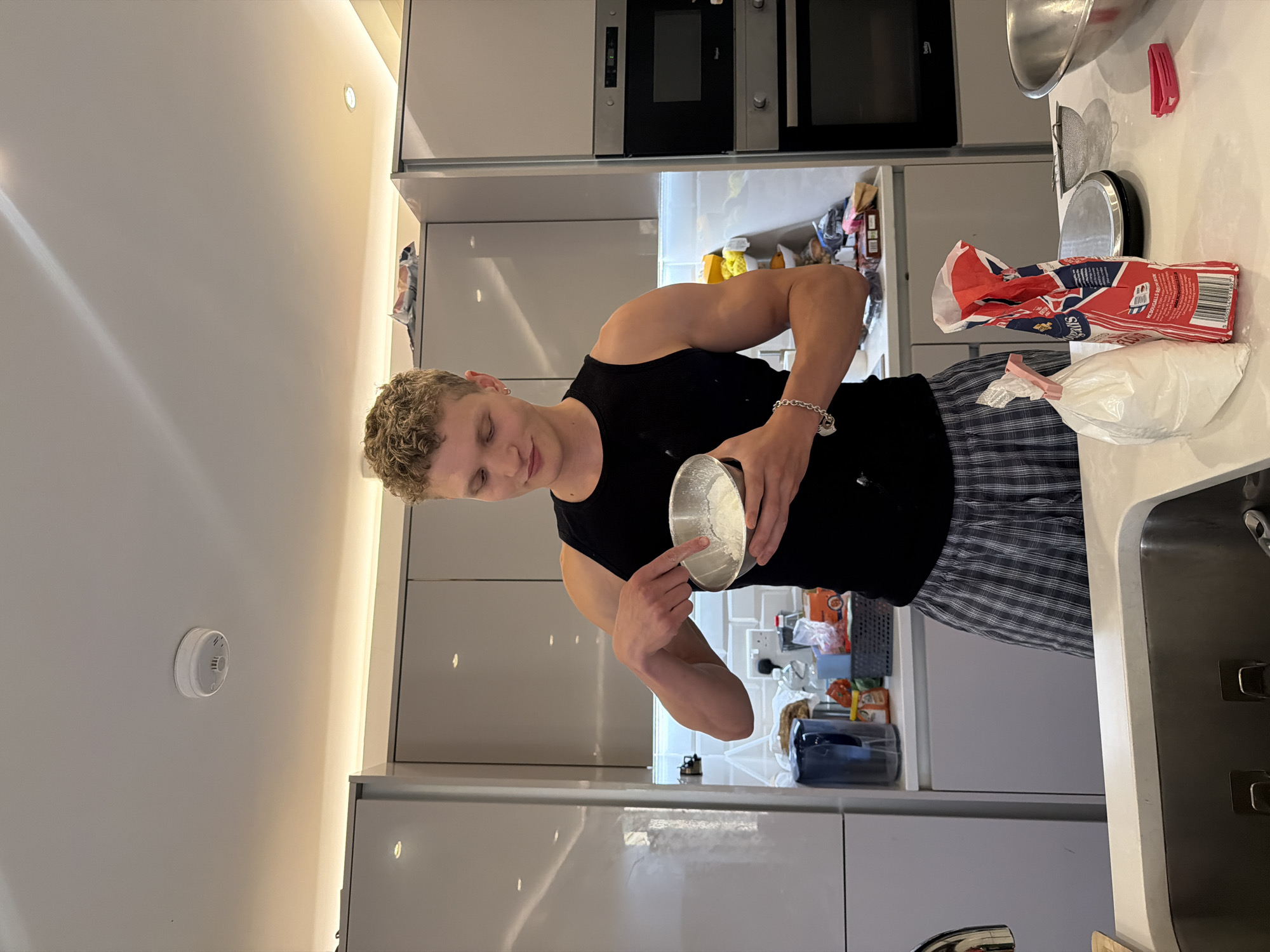 A young man with blond, curly hair and a black tank top is in a modern kitchen, holding a metal bowl filled with a white powdery substance. He is using his index finger to mix the contents of the bowl. He is wearing plaid pajama pants. In the background, there are sleek gray cabinets, a stovetop, an oven, and various kitchen appliances and food items on the counter. A bag of flour and a kitchen scale are also visible on the counter in the foreground. The overall impression is of someone preparing food or ingredients in a home kitchen. The image does not contain enough specific details to determine its location within the city of London, United Kingdom.