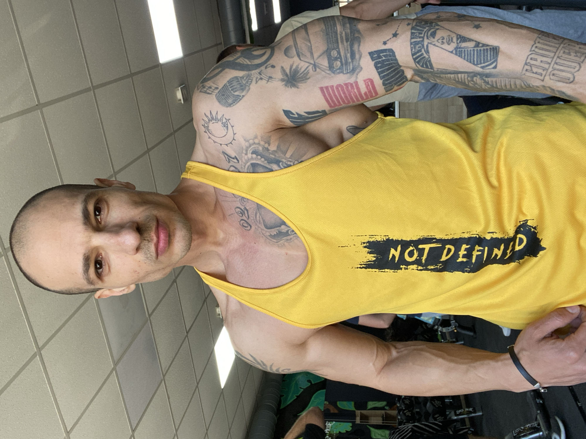 A man with a shaved head and multiple tattoos is posing in what appears to be a gym. He is wearing a yellow tank top with the words "NOT DEFINED" printed on it. The man's skin tone is light brown and he has a short goatee. He is looking directly at the camera with a neutral expression. Tattoos are visible on his arms, shoulders, and chest, featuring various designs including a sun, musical notes, a microphone, and the word "WORLD." Other people are visible in the background, and gym equipment is present. The location is not identifiable as Pleven, Bulgaria. The background consists of a tiled wall and other gym-related elements.