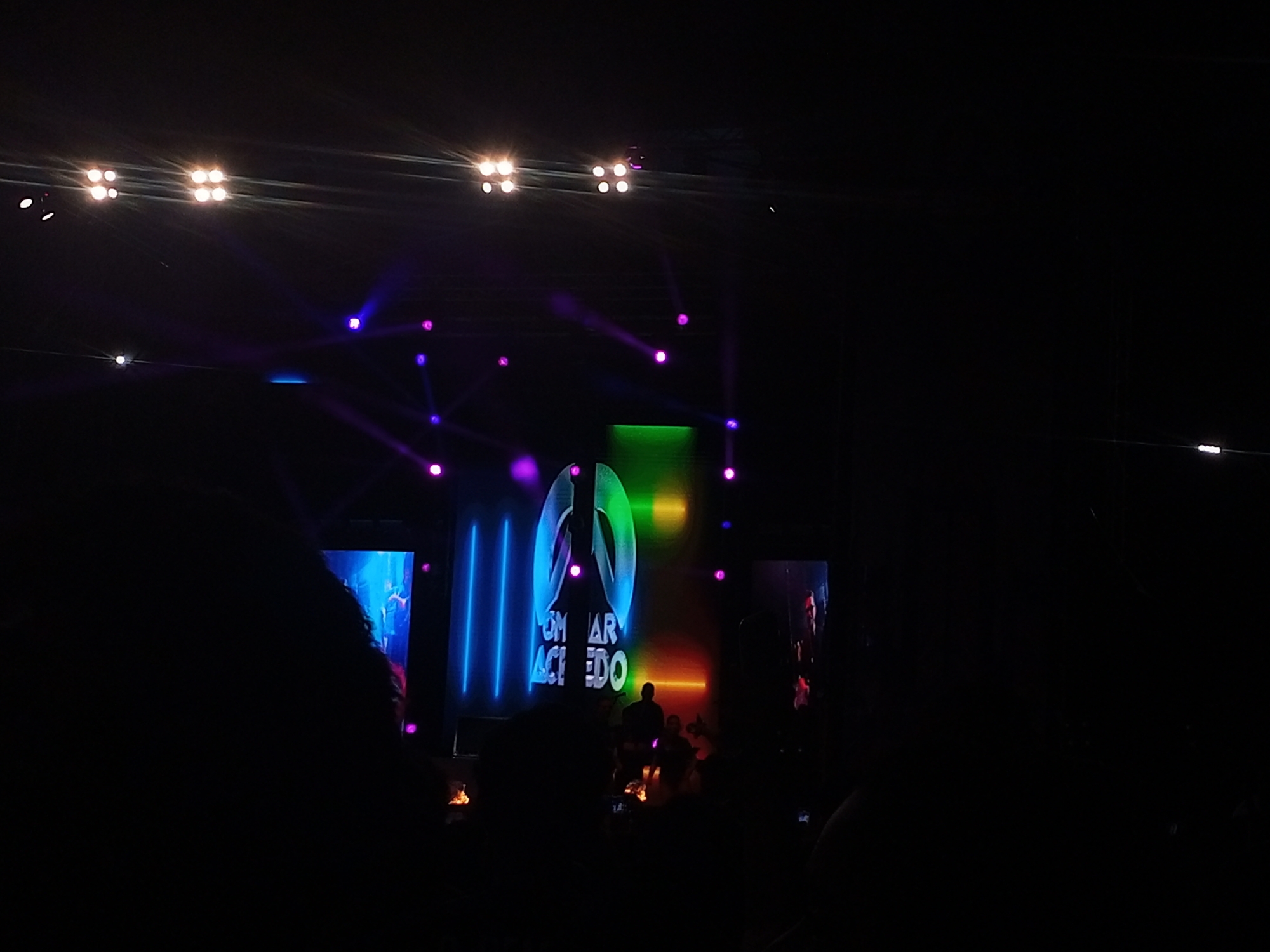 The image depicts a concert or event taking place at night, likely indoors given the controlled lighting. In the background, there is a stage with a large screen displaying the name "OMAR ACEVEDO" along with a logo. The stage is illuminated with various colors, including blue, green, and purple spotlights. There are several people visible on the stage, mostly in silhouette. In the foreground, there is a crowd of people watching the event, their faces mostly obscured by the darkness. Small flames can be seen at stage level.
There is no reliable information in the image to confirm the location of this event in Juan Griego, Venezuela. The description focuses solely on the visual elements present in the image.