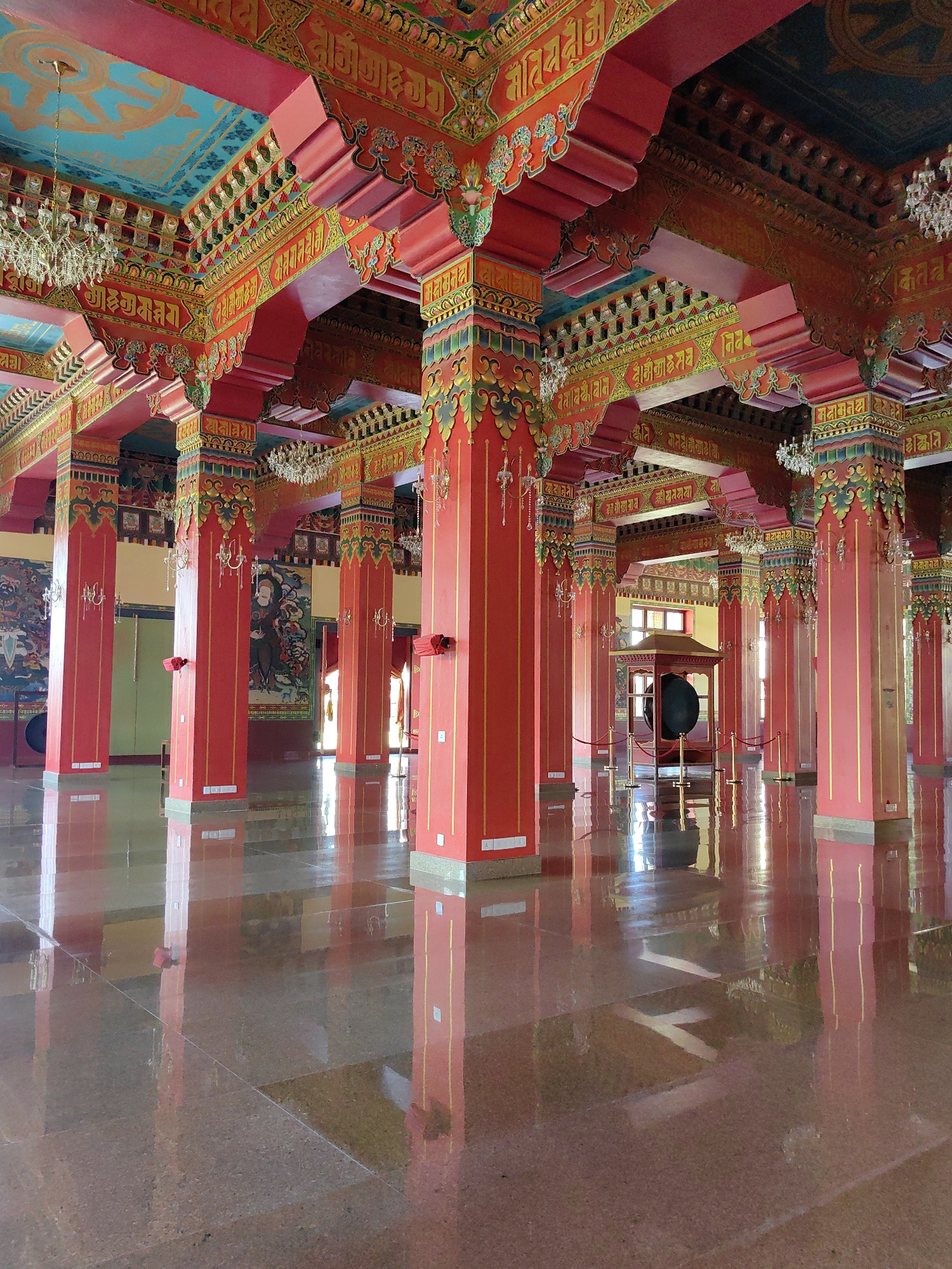 The image depicts the interior of a Buddhist temple, likely the Kagyu Dhargye Ling Monastery, located in Parsa, Nepal. The scene is characterized by vibrant red and golden columns supporting a richly decorated ceiling adorned with Buddhist iconography and script. Chandeliers hang from the ceiling, casting a soft light. The floor is highly polished, reflecting the columns and ceiling decorations, creating a sense of depth and grandeur. In the background, large paintings of Buddhist deities are visible on the walls. A large gong, housed in a protective structure, is centrally located in the middle ground. The overall atmosphere is serene and spiritual, indicative of a sacred space.