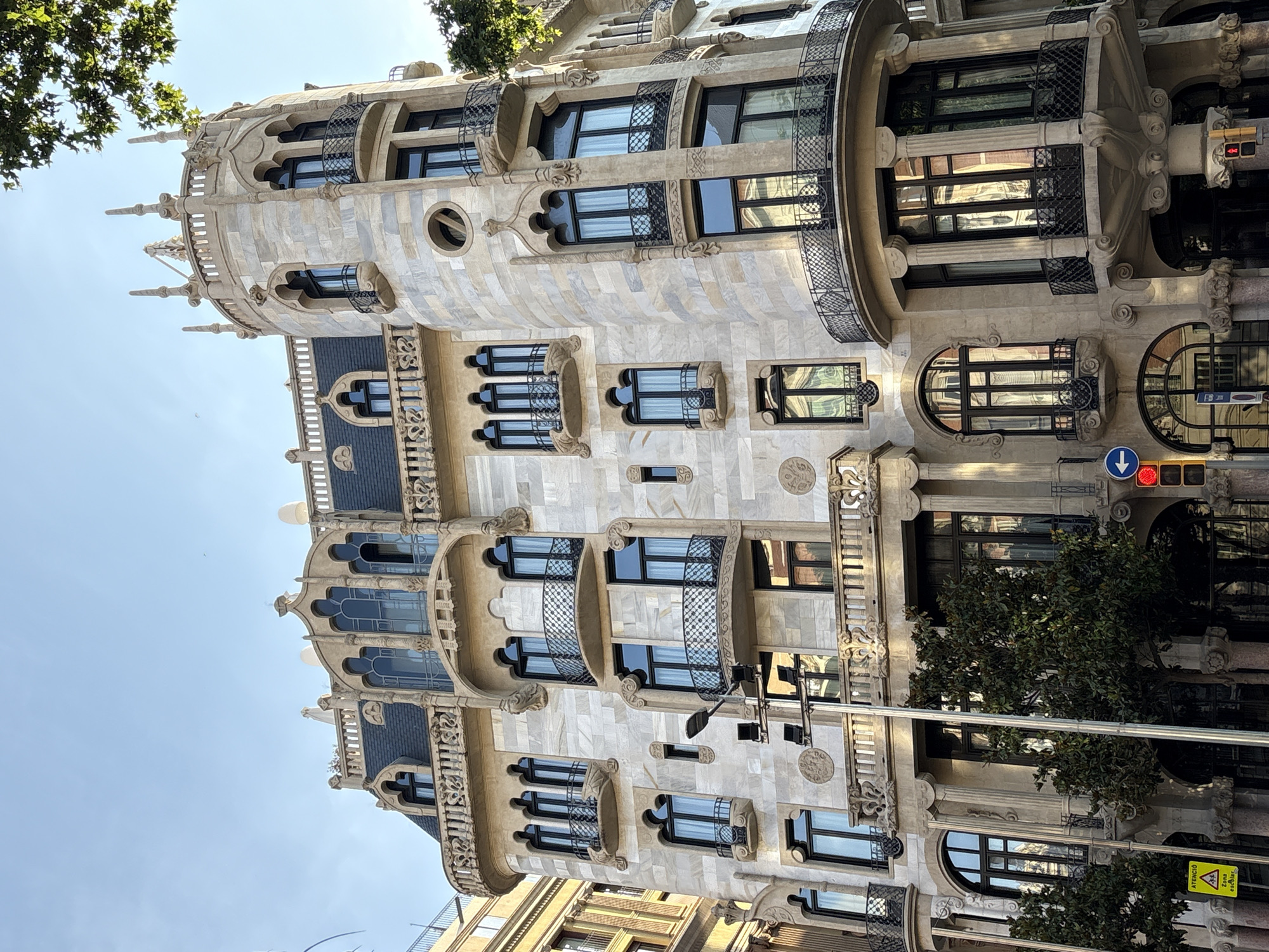 The image captures a portion of the façade of Casa Batlló in Barcelona, Spain, an architectural masterpiece designed by Antoni Gaudí. The building's unique features include irregular window shapes, balconies with wrought-iron railings resembling masks, and an undulating roof. The façade is decorated with colorful mosaic tiles, adding to the building's distinctive character. A traffic light is visible in the scene, indicating that the building is located on a busy street. The sky is blue, and the lighting suggests it is daytime. There are trees and a streetlight in the foreground.