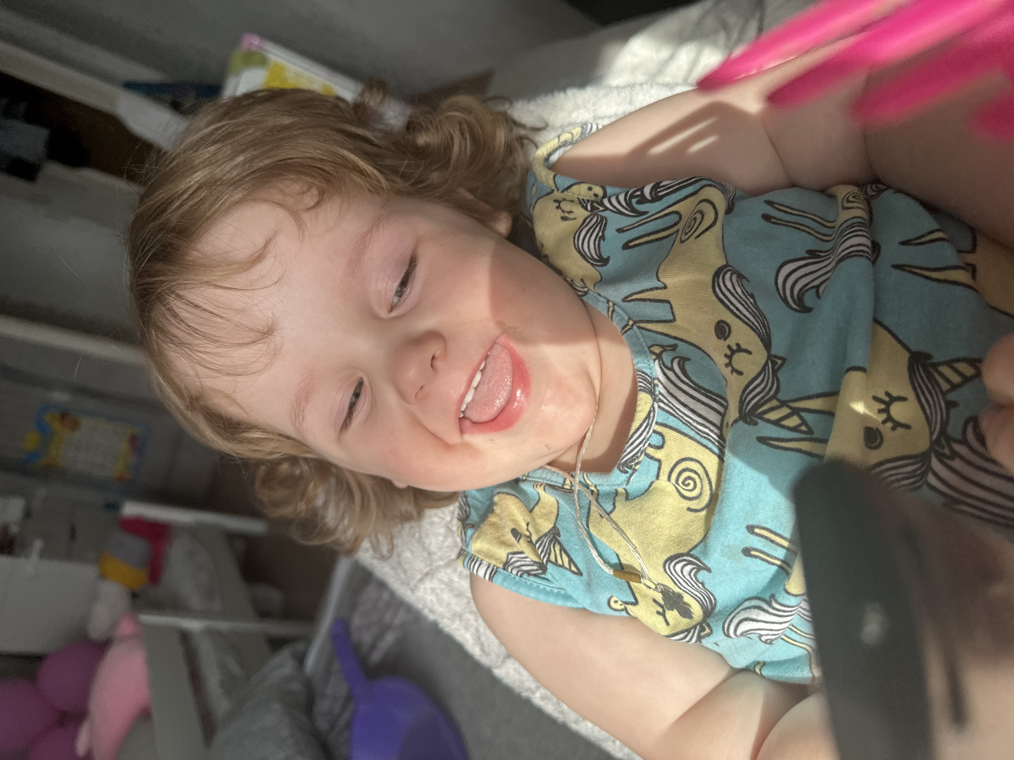 A young child is lying on a light-colored surface, possibly a blanket or cushion. The child has light brown hair and is wearing a blue and yellow shirt with a unicorn pattern. The child's eyes are partially closed, and their tongue is sticking out. A silver necklace with a small brown pendant is visible around the child's neck. In the background, there is a shelf or cabinet with various objects, including toys and books. A purple object sits on the surface near the child. A hand with brightly colored pink nails is partially visible in the top right corner of the frame. The location in the city of Chaikovsky, Russia, is not determinable from the image.