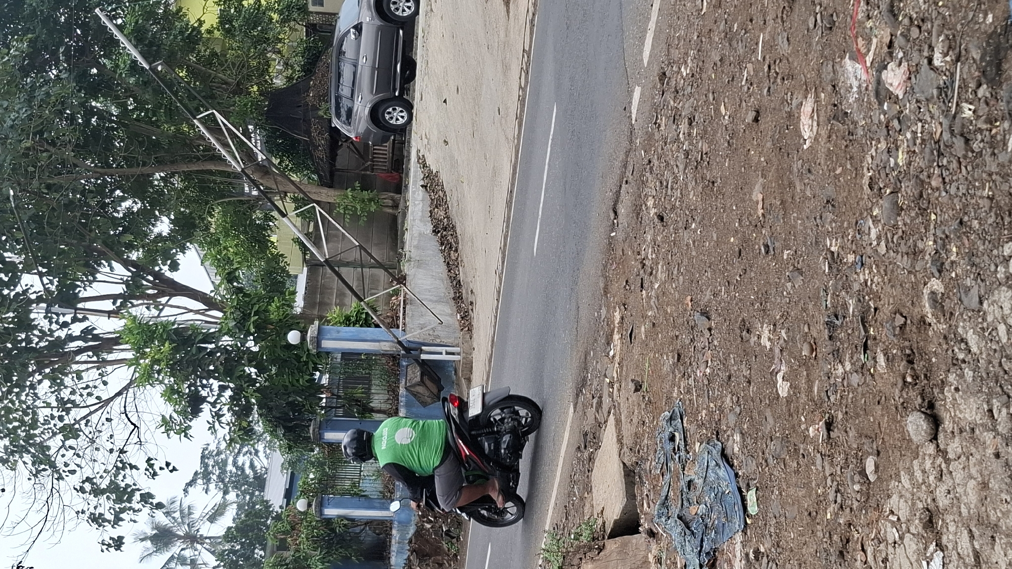 A man wearing a green vest and a black helmet rides a black motorcycle on a paved road in Cilaku, Indonesia. The man is facing away from the camera. The motorcycle has a license plate. The road is bordered by a dirt shoulder and a low concrete barrier. Behind the barrier is a fence and some greenery. In the background, a grey SUV is parked on a light-colored surface. A leaning metal structure, possibly a utility pole or part of a sign, is also visible. The sky is overcast. The environment appears to be rural or semi-urban.