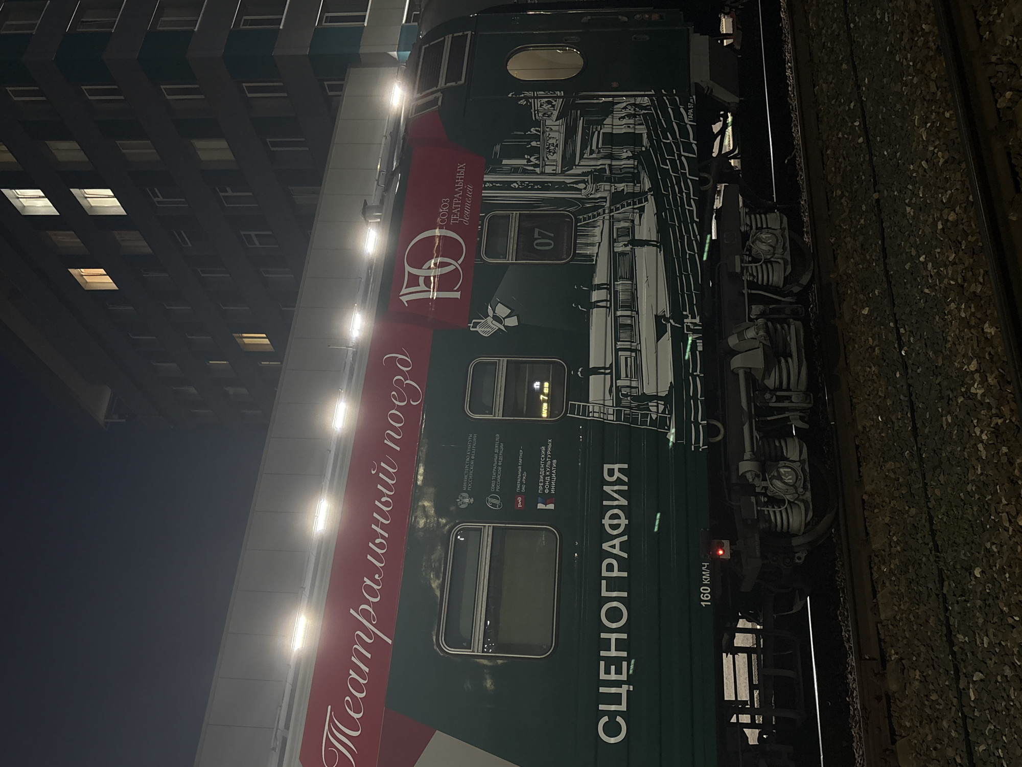 The image captures a side view of a decorated train car at night. The train car is dark green with white lettering and graphics. A red banner stretches across the top of the car with white text that appears to say "Театральный поезд" (Theatrical Train) and a large "150" logo. Below this, the text "СОЮЗ ТЕАТРАЛЬНЫХ ДЕЯТЕЛЕЙ" (Union of Theatre Workers) is visible. The word "СЦЕНОГРАФИЯ" (Scenography) is prominently displayed in large white letters on the lower part of the car. The graphic on the train depicts a theater scene with an audience and stage elements, and the number "07" is visible within a frame. A sign indicating "160 KM/H" is present near the bottom of the car. The train is on tracks, with ballast visible. In the background, a multi-story building with lit windows is dimly visible against the night sky. The overall scene suggests an event or exhibition related to theater, possibly a mobile exhibition or a themed train tour. The location is identified as Ulyanovsk, Russia.