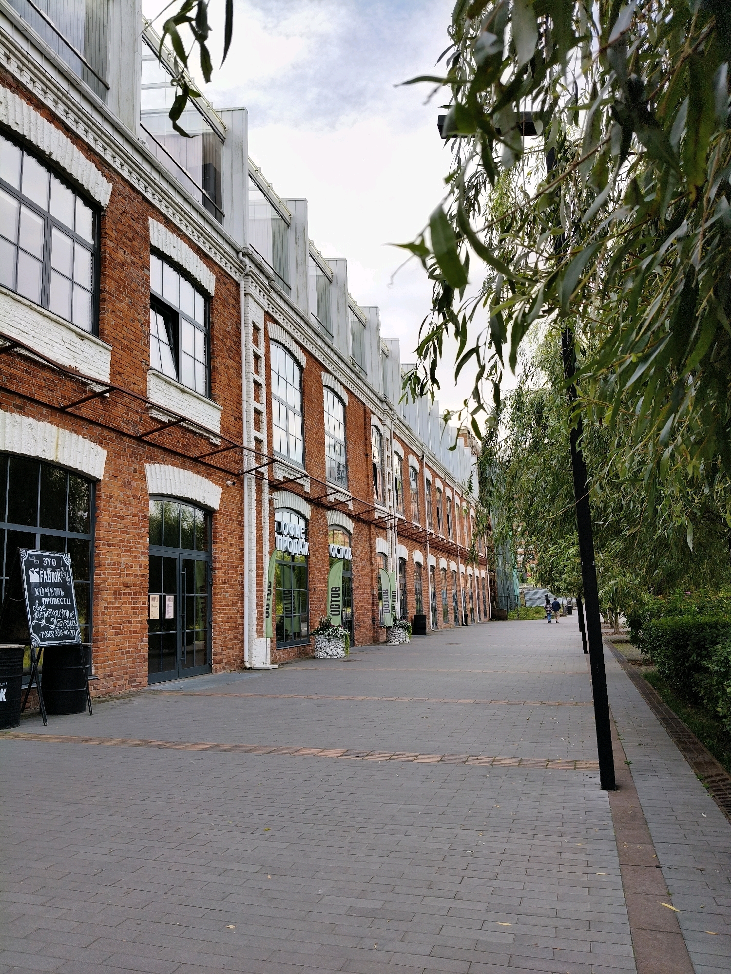 The image shows a brick building with a row of large windows. In front of the building is a paved walkway with a line of trees to the right. A black lamp post stands near the trees. In the distance, two people are walking away from the camera. A blackboard sign is placed in front of the building on the left side of the walkway, with text written on it. The building appears to be an industrial or commercial structure. The overall setting suggests an urban environment, possibly a renovated industrial area.