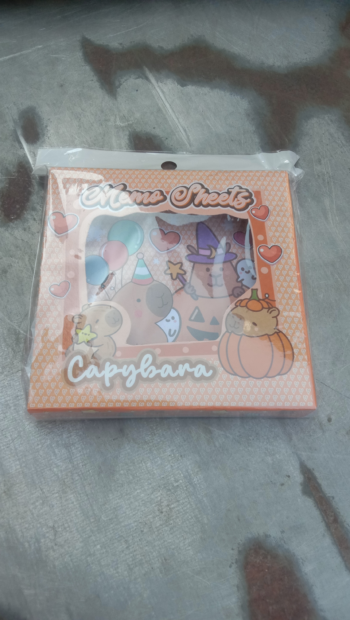 An orange-toned package of memo sheets, likely featuring capybara characters in a Halloween theme, rests on a textured, gray concrete surface. The packaging has a clear plastic wrap, with some visible creasing. The memo sheets themselves display illustrations of capybaras dressed in a witch's hat, sitting on a pumpkin, and other festive elements like balloons and ghosts. The word "Capybara" is printed in large, stylized white lettering on the front of the package. The concrete surface appears somewhat weathered and has dark, damp patches, suggesting it might be outdoors or in an area exposed to moisture. There are no people or animals visible in the image. The overall scene is a close-up, overhead shot of the product.