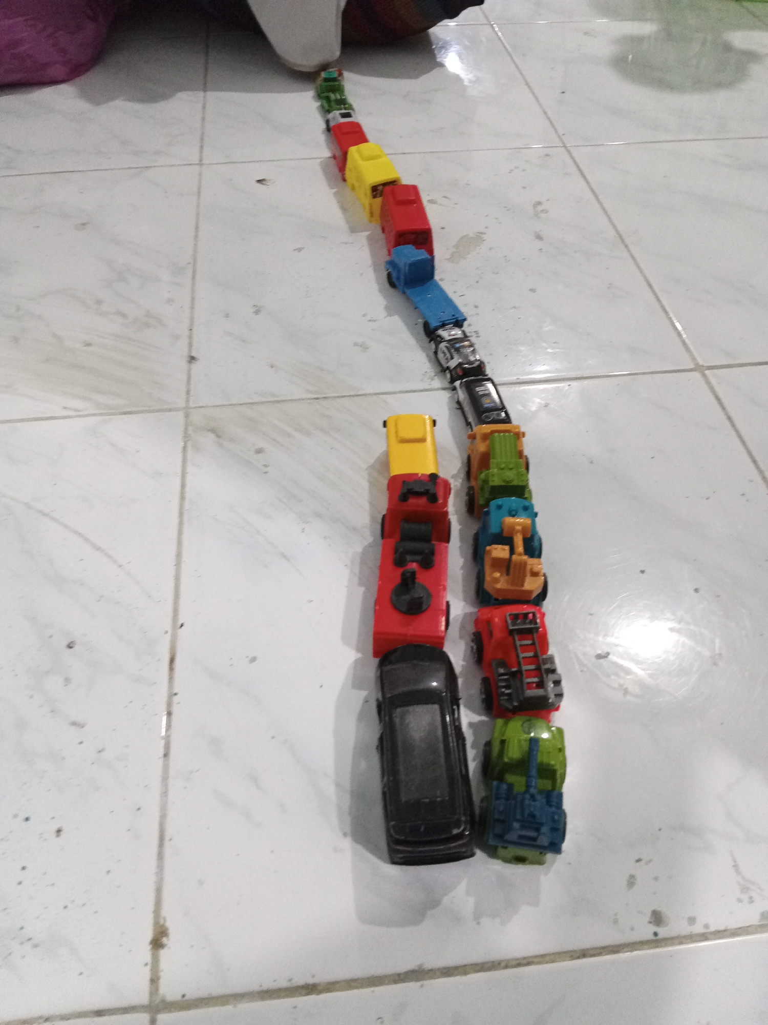 In Lhokseumawe, Indonesia, a collection of toy vehicles are arranged in a line on a tiled floor. The toys, predominantly trucks and cars, are brightly colored in red, yellow, blue, green, and black. The scene is dimly lit, with the tiled floor appearing white and slightly grimy, marked by faint shadows. The toys are positioned diagonally across the frame, with the line beginning near the top right and extending towards the bottom left. The arrangement suggests a deliberate placement, possibly for a game or display. No people are visible in the image.