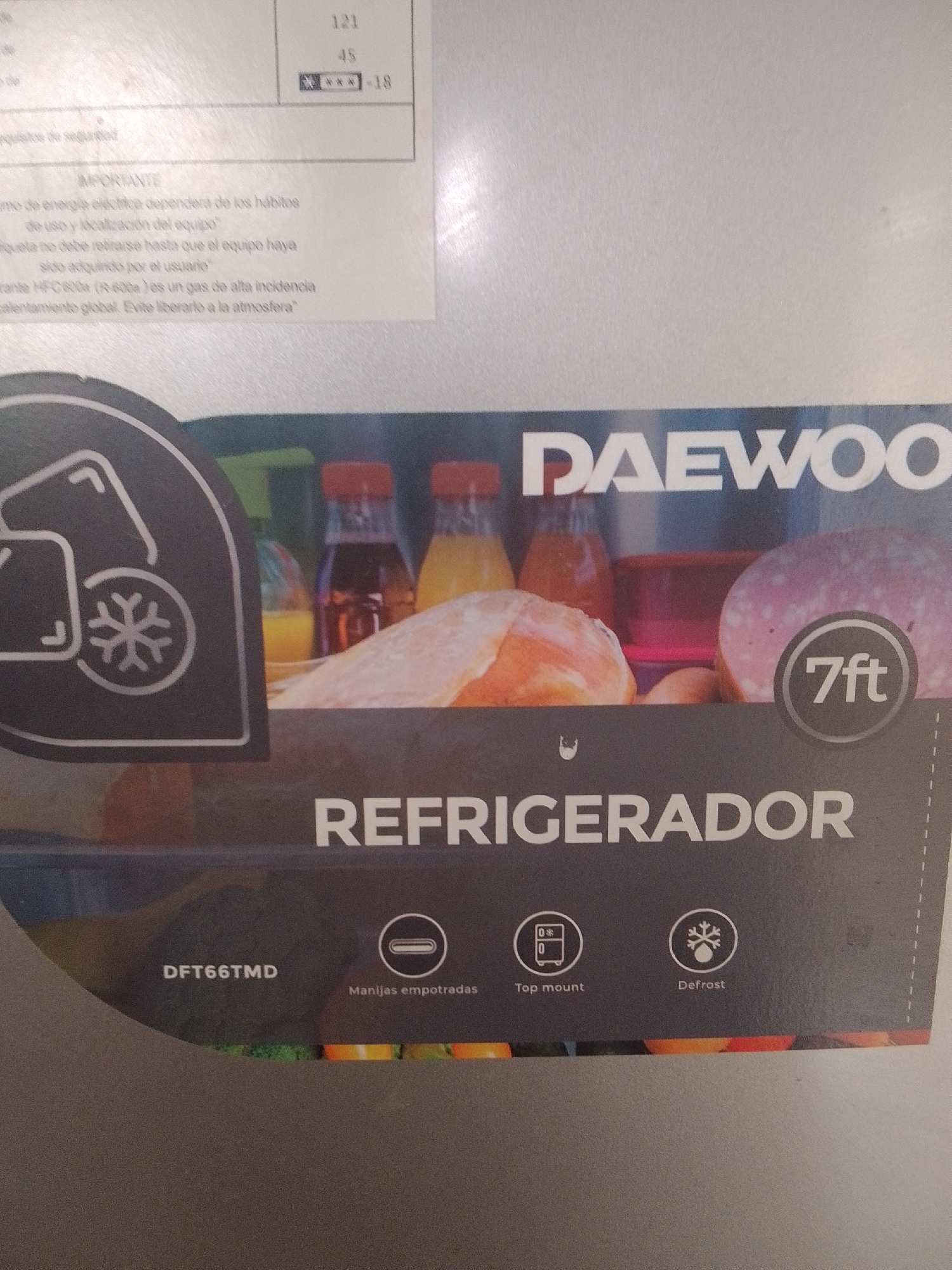 This is a close-up, eye-level, medium shot of a refrigerator. The refrigerator is silver and has a black panel on the bottom half. On the black panel, the brand name "DAEWOO" is written in large white letters. Below the brand name, the word "REFRIGERADOR" is written in even larger white letters. To the right of "REFRIGERADOR", the text "7ft" is visible within a circular emblem.
On the left side of the black panel, the model number "DFT66TMD" is written in white letters. Further to the right, there are three circular icons with text below them. The first icon depicts a refrigerator with "0*" and "0" inside, and the text "Top mount" below it. The second icon shows a snowflake, and the text "Defrost" is below it. The third icon shows a set of handles, with the text "Manijas empotradas" below it.
The upper portion of the image shows a label with some text in Spanish. It discusses important information regarding energy consumption and the environmental impact of the refrigerant used. There are also some numbers visible, including "121" and "45", along with a rating of "***-18". The background of the upper portion of the image is blurred but appears to be a wall or a similar surface. The overall impression is that this is an informational label or a product image. The context provided is La Tagua, Venezuela, suggesting this is a product sold or used in that location. There are no people visible in the image.