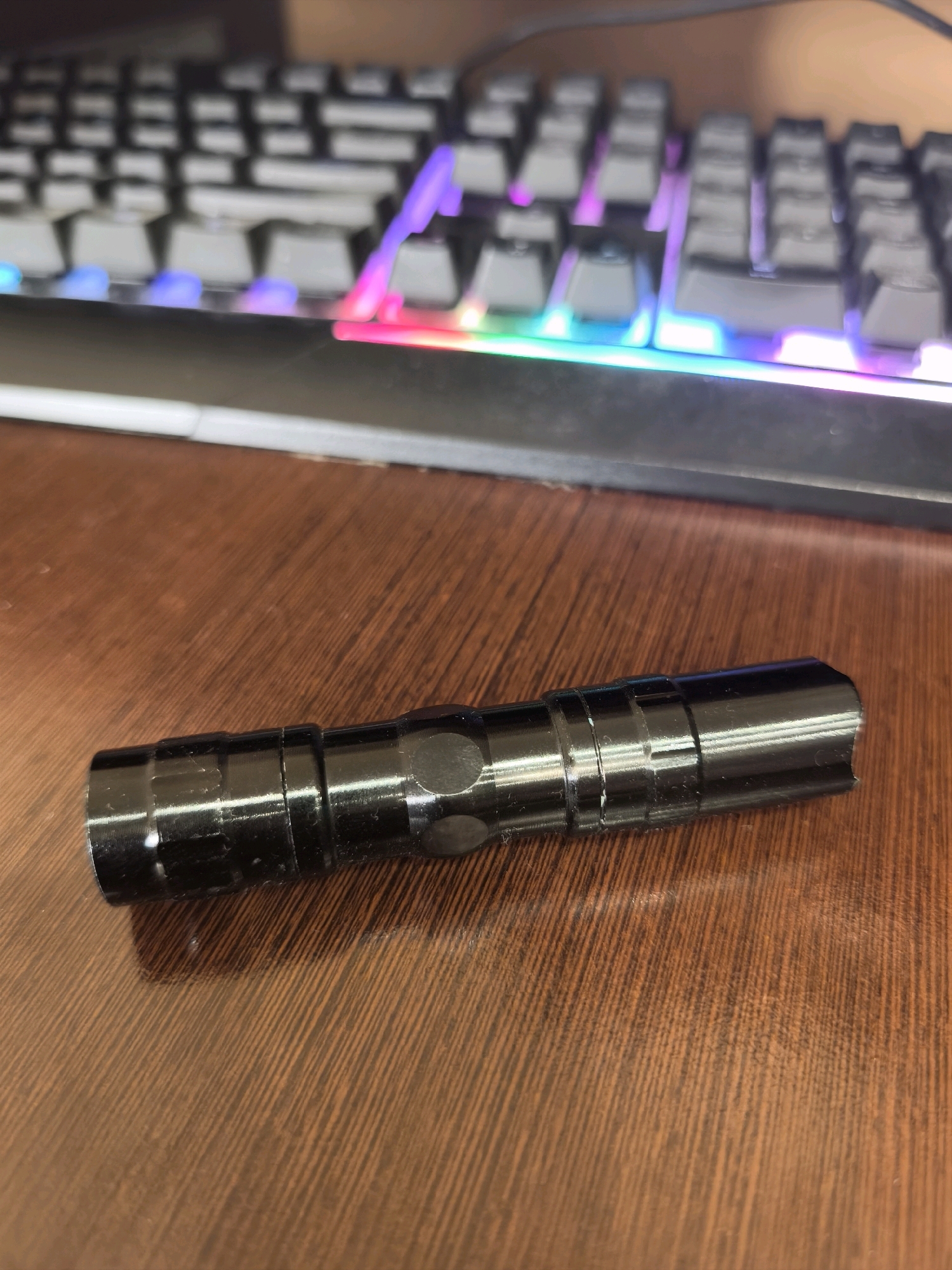 A black metal flashlight lies on a wooden desk in front of a blurred RGB gaming keyboard. The flashlight has a ribbed body with several distinct sections. The desk has a visible wood grain pattern. The keyboard has black keys and is illuminated with colorful lights in shades of purple, blue, and pink. The image does not contain any information to determine its location in Karachayevsk, Russia.