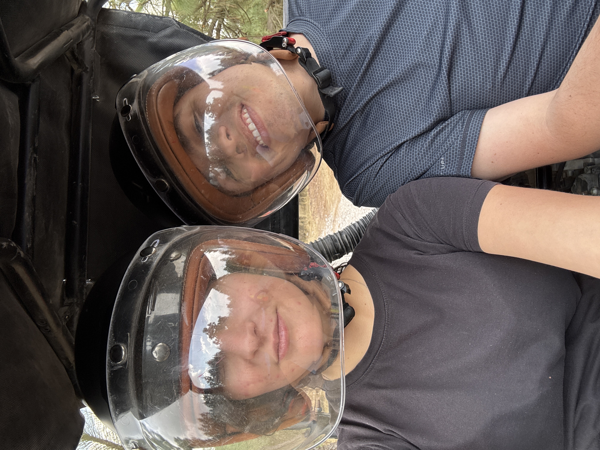 Two individuals wearing clear helmets stand close together. One person is wearing a dark blue patterned shirt, while the other wears a black t-shirt. They appear to be in a natural setting with trees visible in the background. The location is likely San Juan Arcos Ojo de Agua, Mexico.