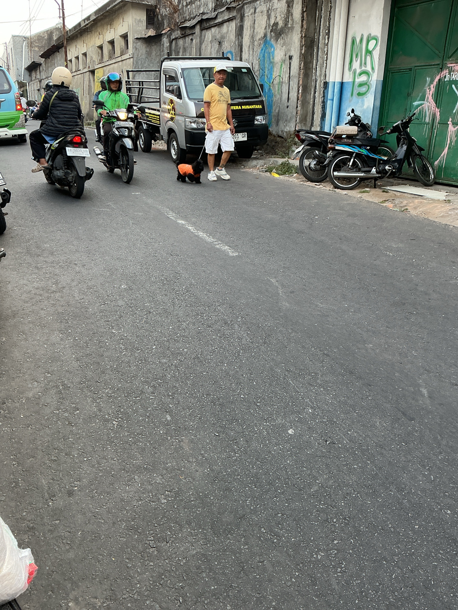 On a street in Magelang, Indonesia, a man walks his black dog, which wears an orange jacket. The man is wearing a yellow t-shirt, white shorts, a yellow cap, and white shoes. He is holding the dog's leash. Behind him is a silver truck with "JURUNUSA NUSANTARA" written on the side. To the left of the truck, two people are riding a motorcycle. The driver of the motorcycle is wearing a green jacket, and the passenger is wearing a black jacket and a beige helmet. In front of them, another person is riding a motorcycle while wearing a black jacket and a beige helmet. Parked along a wall with green doors are three motorcycles. The wall has graffiti on it, including "MR 13" in green. The street is paved, and there are buildings on the left side of the image. The sky is overcast.