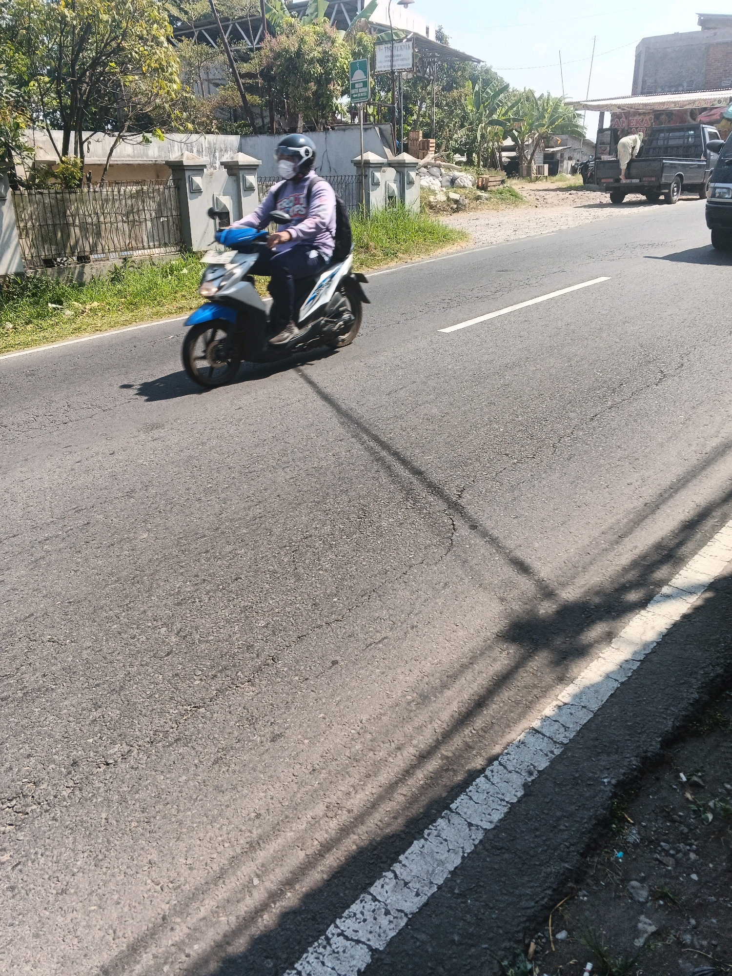 The image captures a street scene in Bandungan, Indonesia, under a bright, clear sky. A person wearing a light purple long-sleeved shirt, dark pants, a dark helmet, and a white mask is riding a white and blue scooter. The scooter's front wheel is blurred due to motion. In the background, there is a wall with ornate pillars and a metal fence, beyond which are trees and some buildings. A signpost is visible on the right side of the road, with text that appears to be in Indonesian. Further in the background, a dark-colored pickup truck is parked, with a person standing in its bed. The road is asphalt with a white line marking the edge. Shadows of overhead wires or branches are cast across the road. The environment appears to be a semi-urban or rural setting, with vegetation and structures typical of Indonesian towns.