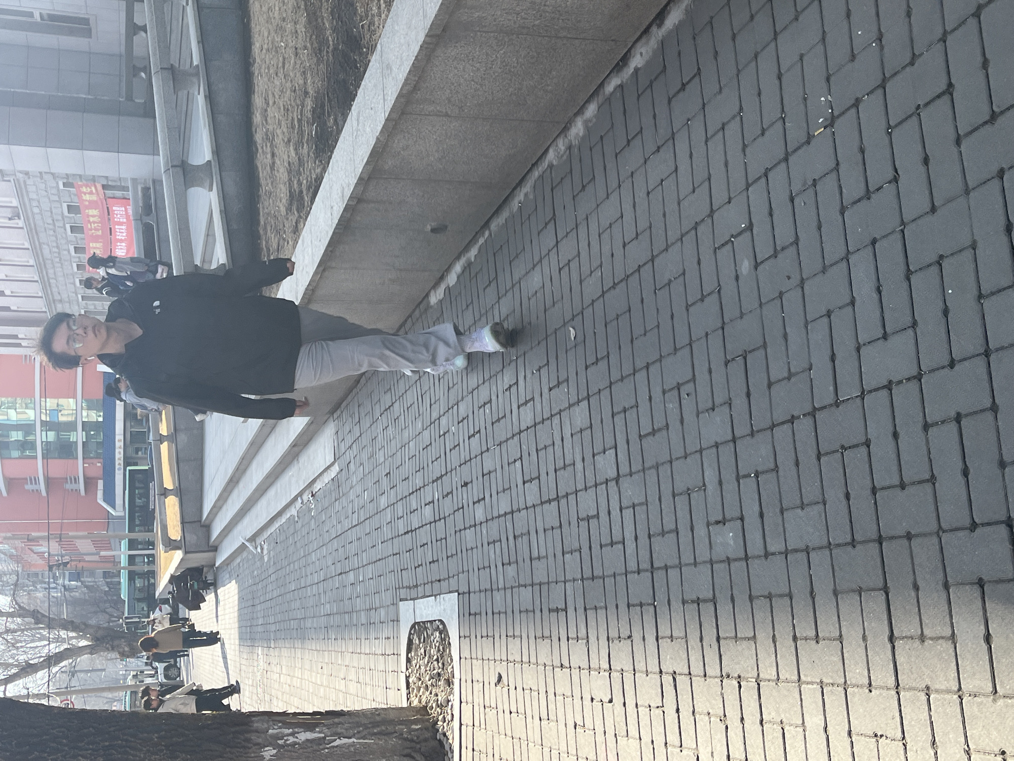 A man wearing glasses, a dark jacket, and gray pants stands on a paved sidewalk in Changchun City, China. The sidewalk is constructed of rectangular gray bricks. A concrete planter or retaining wall runs along the left side of the sidewalk. In the background are buildings, including a tall one with red and white colors and a sign with Chinese characters. Other people are walking further in the background near a bus stop. Bare trees and some snow are visible near the bottom of the frame. A small, circular or oval area filled with pebbles or small stones is embedded in the sidewalk near the bottom of the image.