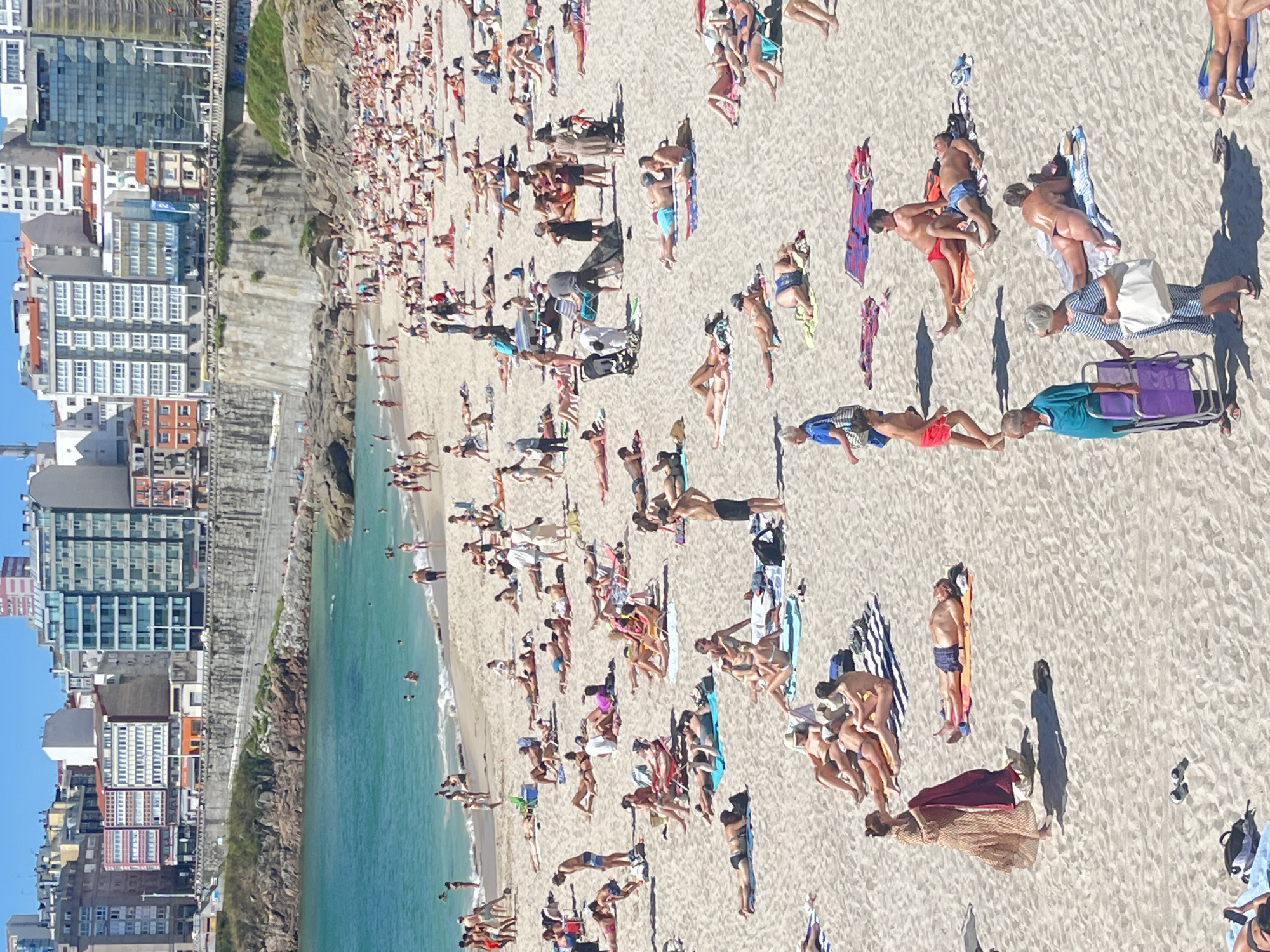This aerial photograph depicts a crowded beach in A Coruña, Spain. Numerous people are sunbathing on the sand, scattered across the scene. Some individuals are lying on towels, while others are seated or standing. The beach is adjacent to the ocean, with gentle waves visible near the shore. Buildings, including a high-rise apartment complex and other structures, are visible along the coastline, suggesting an urban beach setting. The scene is bathed in bright sunlight, indicating a clear day, likely during the warmer months given the attire of the people. The overall atmosphere appears relaxed, as people are engaged in leisure activities. There is no visible text in the image.