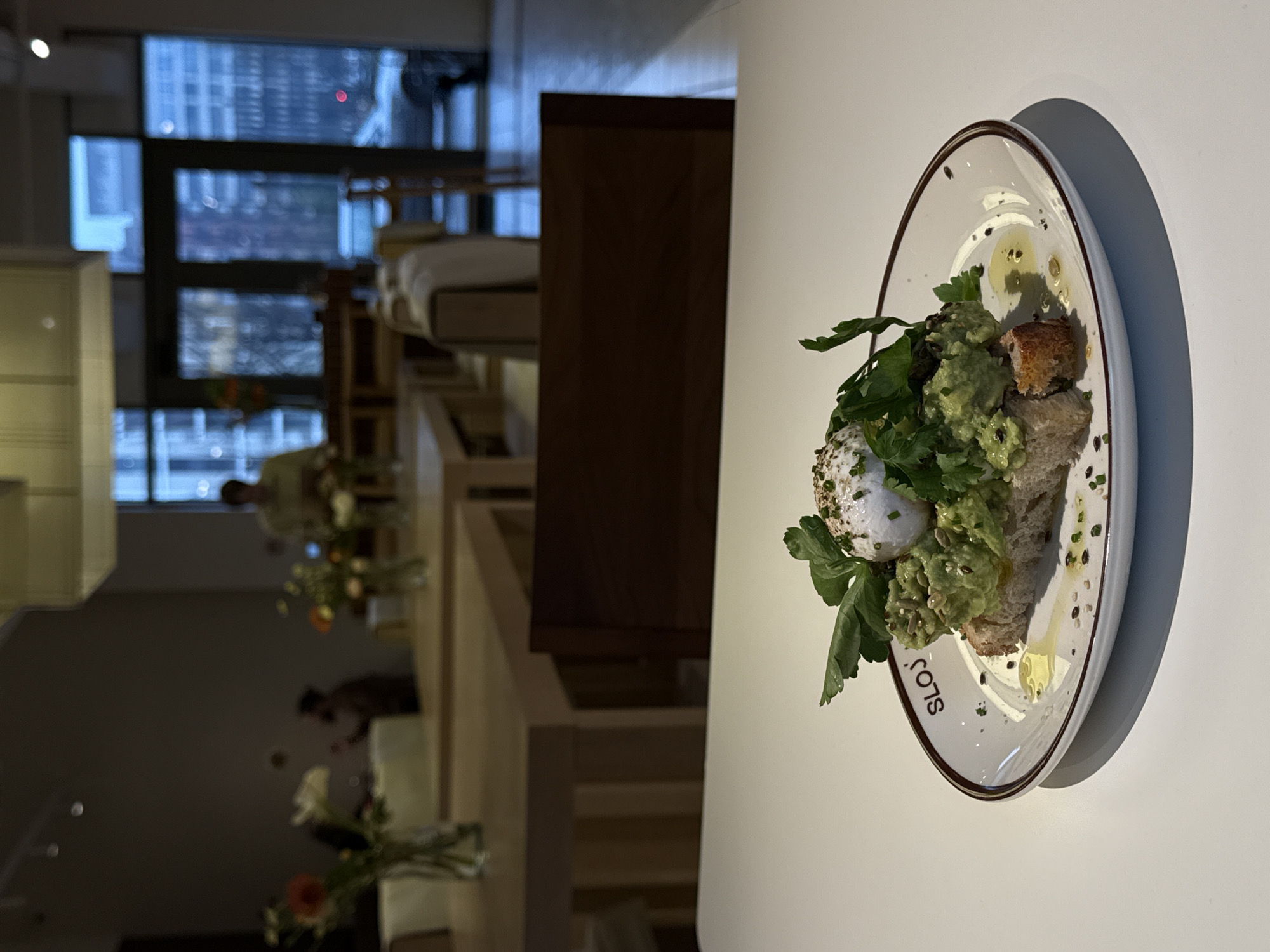 The image shows a plate of avocado toast with a poached egg and herbs on a white table. The plate has a brown rim and the word "SLOJ" written on it, which suggests the dish is from the SLOJ restaurant. The background shows a blurred interior of a restaurant with a view of buildings and a cityscape through the windows. Based on the style of the restaurant and the cityscape visible, it is plausible that the location is in the city of Belgrade, Serbia. The photo focuses on the plate of food, highlighting the ingredients and presentation of the dish. In the background, there is an indication of other people and tables, but the main subject is clearly the food.