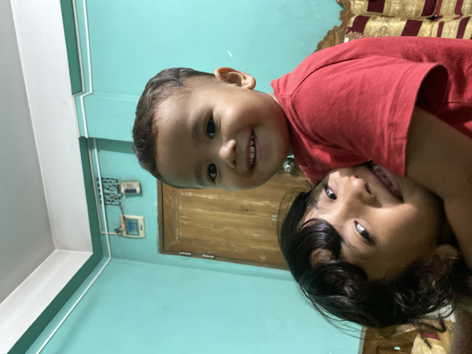 Two children are positioned closely together in an indoor setting. One, wearing a red shirt, is in the background, while the other, with black hair, is closer to the camera. Both are smiling. The background includes a turquoise wall, a wooden door, and a section of furniture with a patterned red and gold fabric. Electrical fixtures are visible on the wall. The location is Dhaka, Bangladesh.