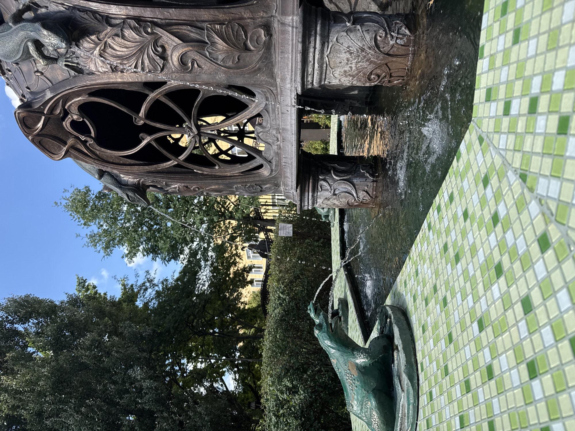 In Sovkhoza imeni Lenina, Russia, a bronze fountain with decorative lizards spouts water. In the foreground, a green tiled area with a mosaic pattern of light and dark green squares leads to a pool. A green frog sculpture sits on the tiled edge, with water spraying from its mouth. The pool is surrounded by lush green bushes and trees, with a yellow building visible in the background under a clear blue sky with scattered clouds.