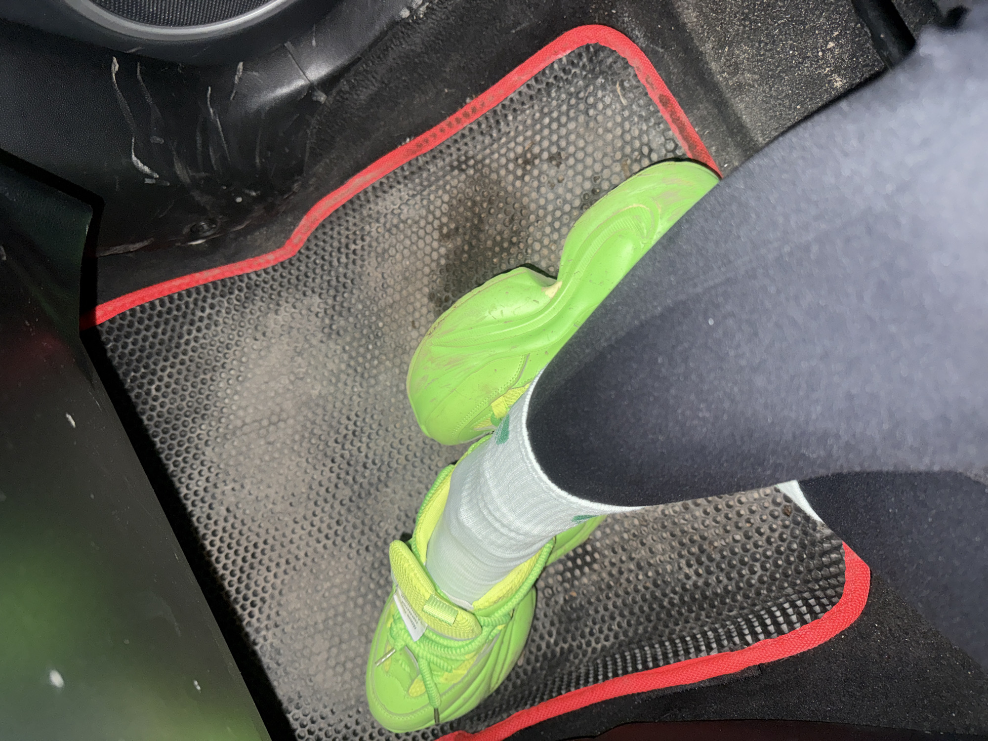 A person wearing a black legging, a white sock with green stripes, and a neon green sneaker is resting their foot on a black rubberized car floor mat with a red trim. The floor mat shows signs of wear and dirt. Part of the car's interior, including a section of the dashboard and the lower portion of a door, is visible. The exact location within Kauchuk Stantsiya, Russia, cannot be determined from the image.