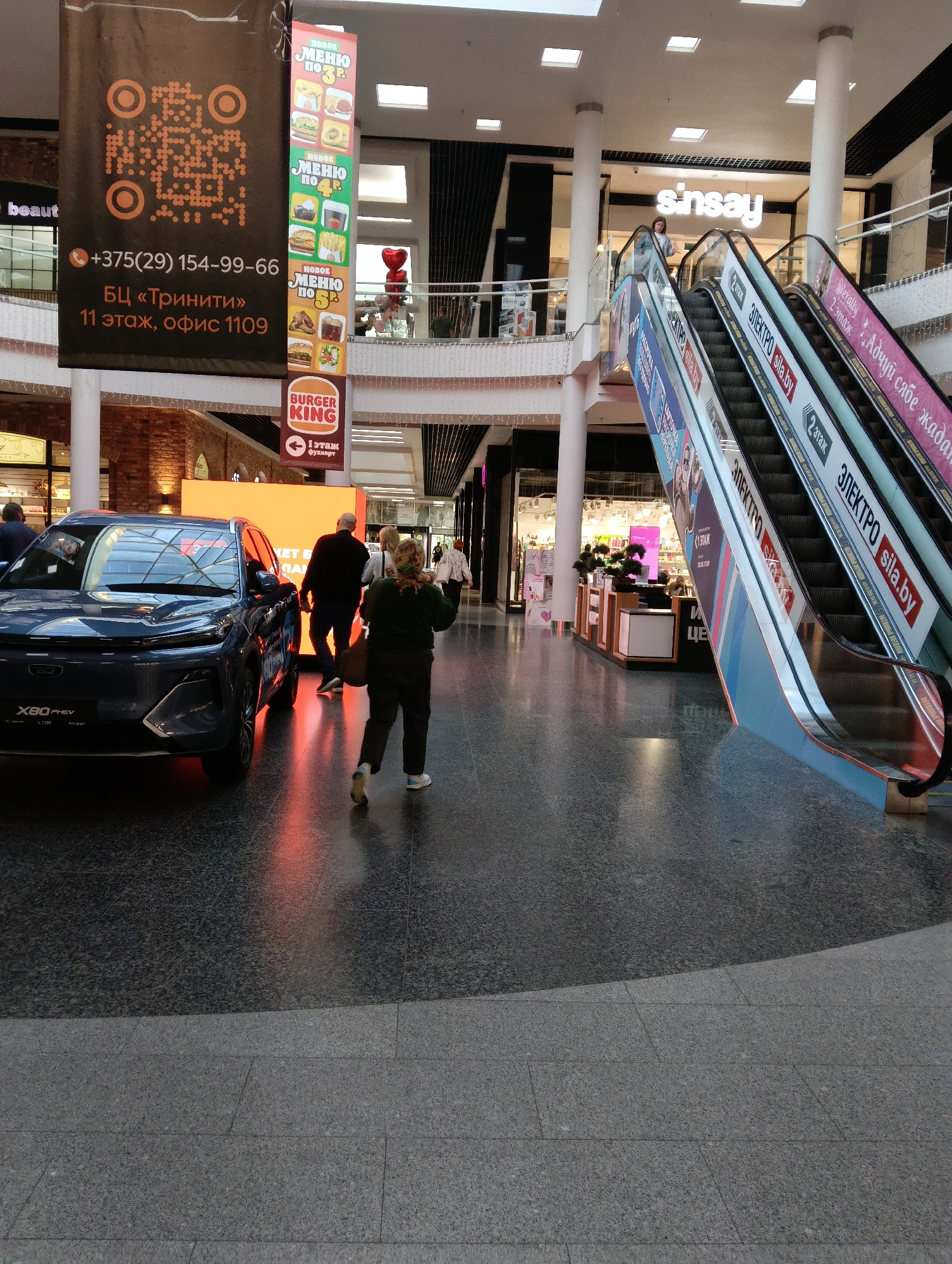 This image shows an interior view of a shopping mall. In the foreground, a dark blue car, identified by the "X80 PHEV" badging, is parked on the left. Behind the car, several people are walking. To the right of the car, a woman in a green jacket and dark pants is walking away from the camera. Further back, two men and another woman are walking in the same direction.
Dominating the left side of the image is a large black banner with orange text and a QR code. Text on the banner includes a phone number, "+375(29) 154-99-66", and location details: "БЦ «Триннити» 11 этаж, офис 1109". Another smaller banner below it advertises "BURGER KING".
On the right side of the image, a large escalator with advertisements running along its sides is visible. One advertisement clearly shows the text "sinsay" in large white letters, indicating a store in the mall. Other advertisements on the escalator include Russian text such as "ЭЛЕКТРО sila.by".
The mall has a high ceiling with recessed lighting and decorative elements. The floor is made of dark, speckled material. The overall lighting suggests it is daytime, with natural light filtering in from upper levels or skylights, supplemented by artificial mall lighting. This appears to be a modern commercial interior.