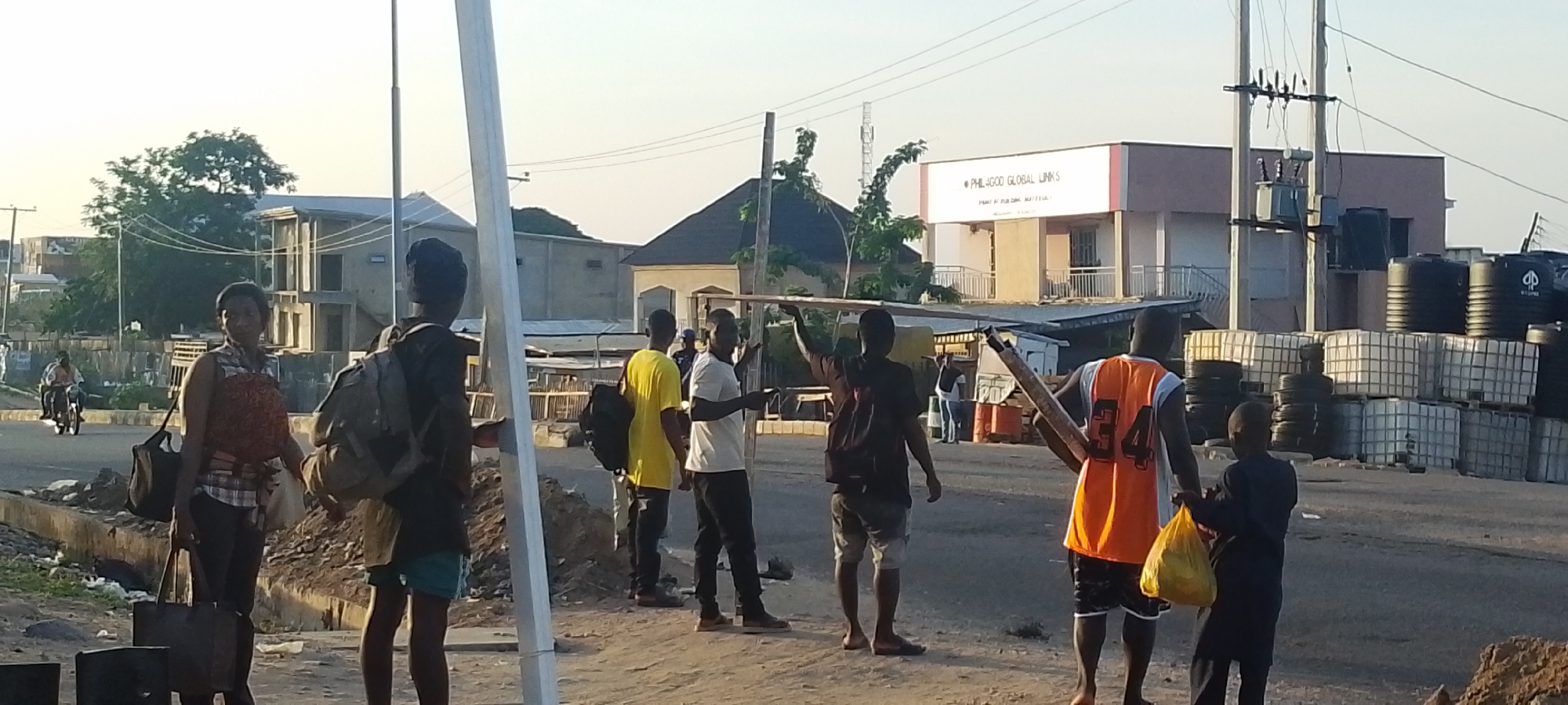 The image depicts a street scene in Lokoja, Nigeria. Several individuals are standing along the roadside. A woman in patterned top and dark pants carrying bags stands to the left of the frame. Nearby, several men are visible, one carrying a backpack and wearing shorts. Several others are gathered, some carrying metal poles. One man is wearing a yellow shirt, and another is in a white shirt. One man is wearing an orange vest with the number 34. A child is present to the right, carrying a yellow bag. In the background, buildings and an overhead power line are visible. The business 'PHIL4GOOD GLOBAL LINKS' can be seen on one of the buildings. Stacks of large water tanks and caged containers are visible behind the group. A motorcycle rider is visible on the road in the distance.