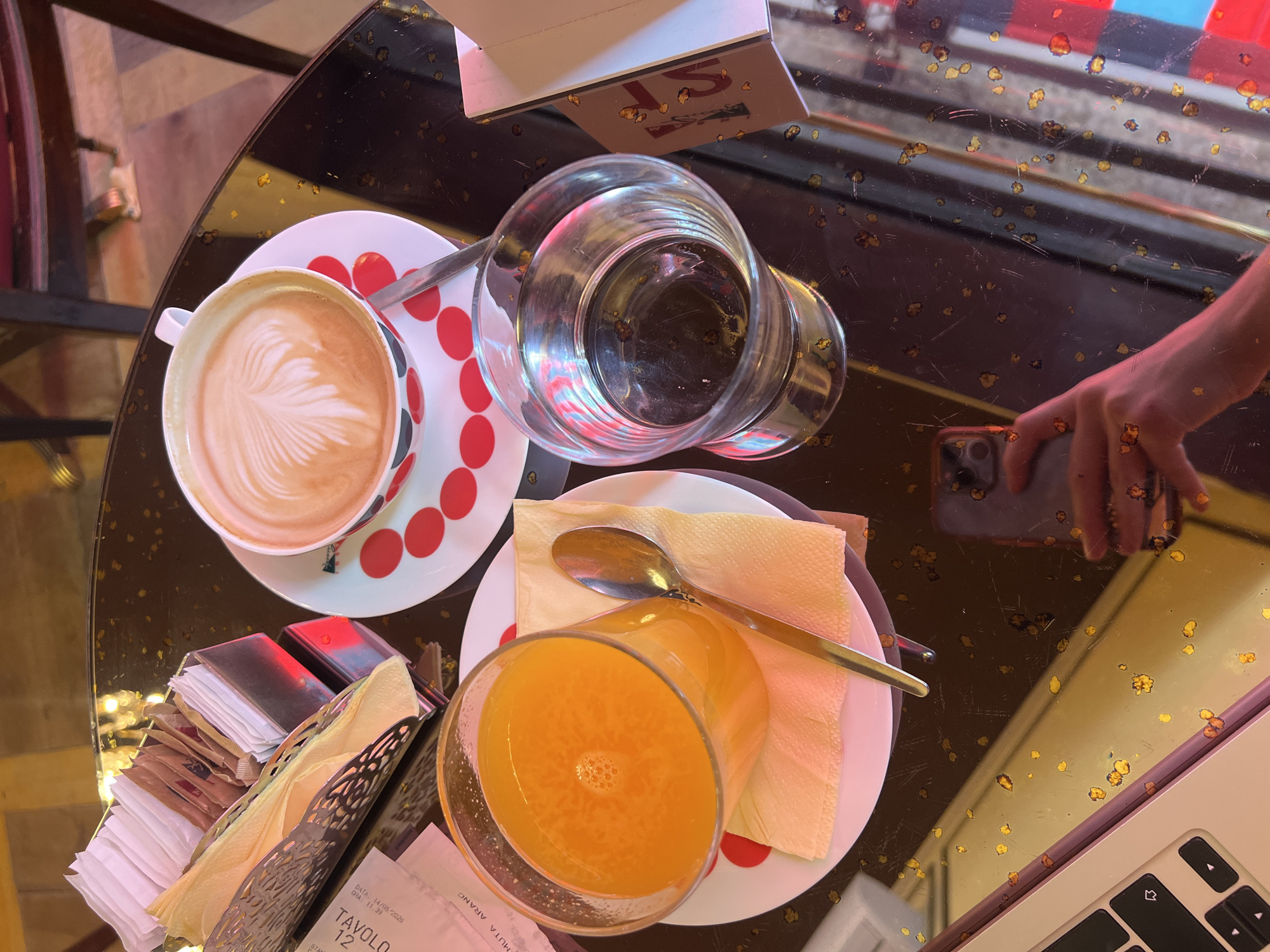 The image depicts a table setting in what appears to be a cafe or restaurant, possibly in Milan, Italy, given the "ARANC" on a receipt. A cup of coffee with latte art sits beside a glass of water and a glass of orange juice, each on matching red-and-white dotted plates. A gold-colored spoon rests on a napkin beside the orange juice. A sugar packet holder with napkins is also on the table. A hand holding a smartphone is seen reflected in the glossy table surface, suggesting someone is taking a photo. Part of a laptop is visible on the edge of the table. The table surface itself has a slightly dirty or aged appearance.