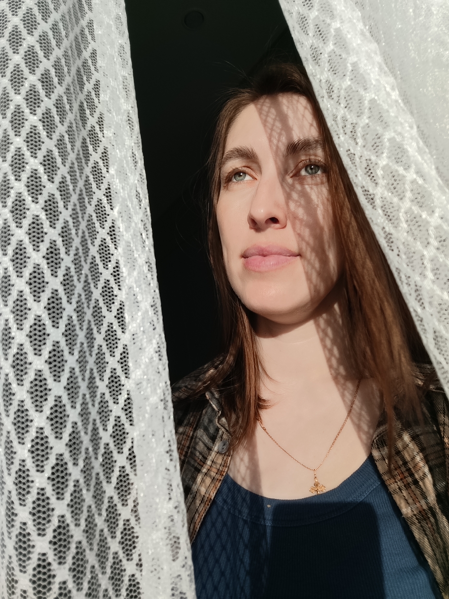 A single female individual is depicted indoors in Chaikovsky, Russia. The subject, appearing to be in her late twenties to mid-thirties, has light skin, light blue eyes, and light brown hair. She is dressed in a dark blue ribbed tank top under an open plaid shirt featuring brown, beige, and dark blue tones. A thin gold chain necklace with a small gold pendant is visible on her chest. She looks upwards and slightly to the viewer's right with a neutral expression. Strong sunlight from the left casts a distinct shadow pattern, consistent with a hexagonal mesh curtain, across her face, neck, and upper torso. She is positioned between two panels of white, translucent curtains, each displaying the aforementioned hexagonal mesh pattern. The background on the right side of the image is dark.