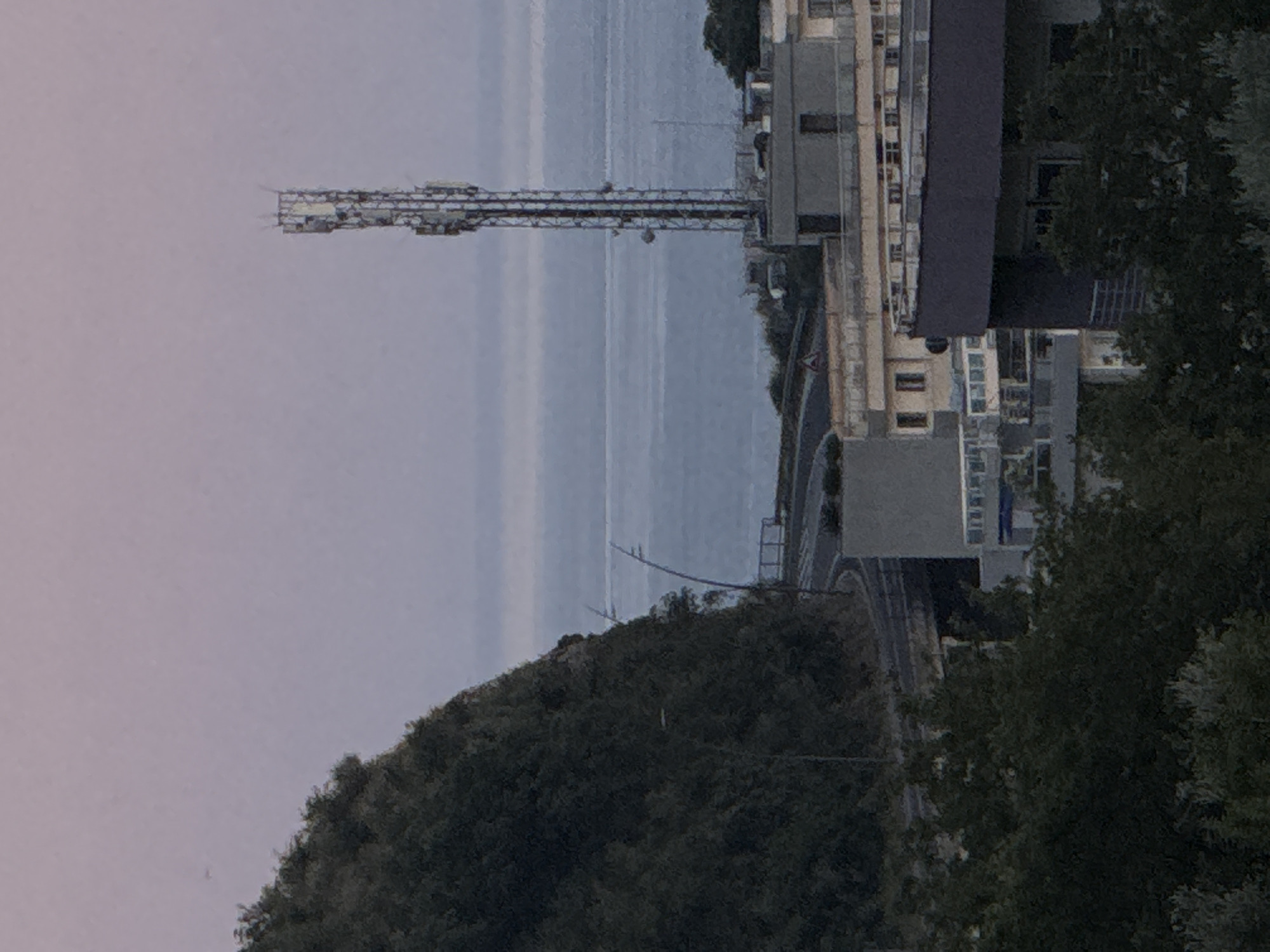 A distant, slightly out-of-focus view of a coastal town, likely Rafailovići, Montenegro, is captured from an elevated perspective at dusk. The scene is dominated by a lush, green hillside on the left, sloping down towards a winding road. Beyond the hillside and road, the calm, hazy blue sea stretches to the horizon, meeting a pale, pinkish-purple sky. A tall, lattice-like telecommunications tower stands prominently against the sky, positioned to the right of the hillside. In the middle ground, several buildings of varying heights and architectural styles are visible, with some featuring balconies and windows. The buildings appear to be a mix of residential and commercial structures. The overall impression is one of a quiet, late evening in a seaside urban area.