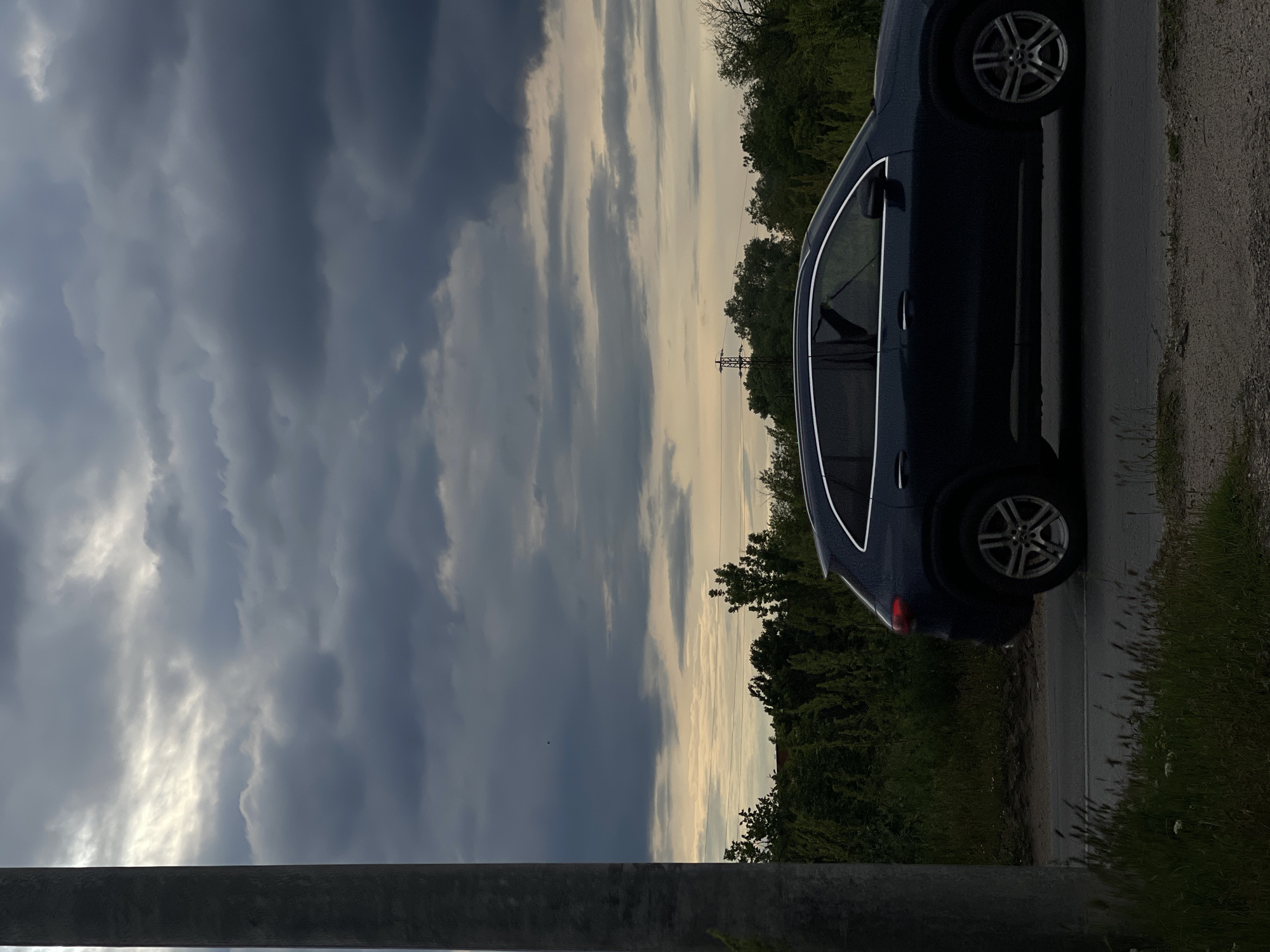 The image shows a dark blue car parked on the side of a road with overgrown vegetation. The sky is filled with heavy, dark clouds, suggesting an approaching storm. In the background, there are trees and an electrical power line tower. A gray pole is visible in the lower part of the image. Based on the vegetation and architecture, it is impossible to confirm that this image was taken in Sofia, Bulgaria.