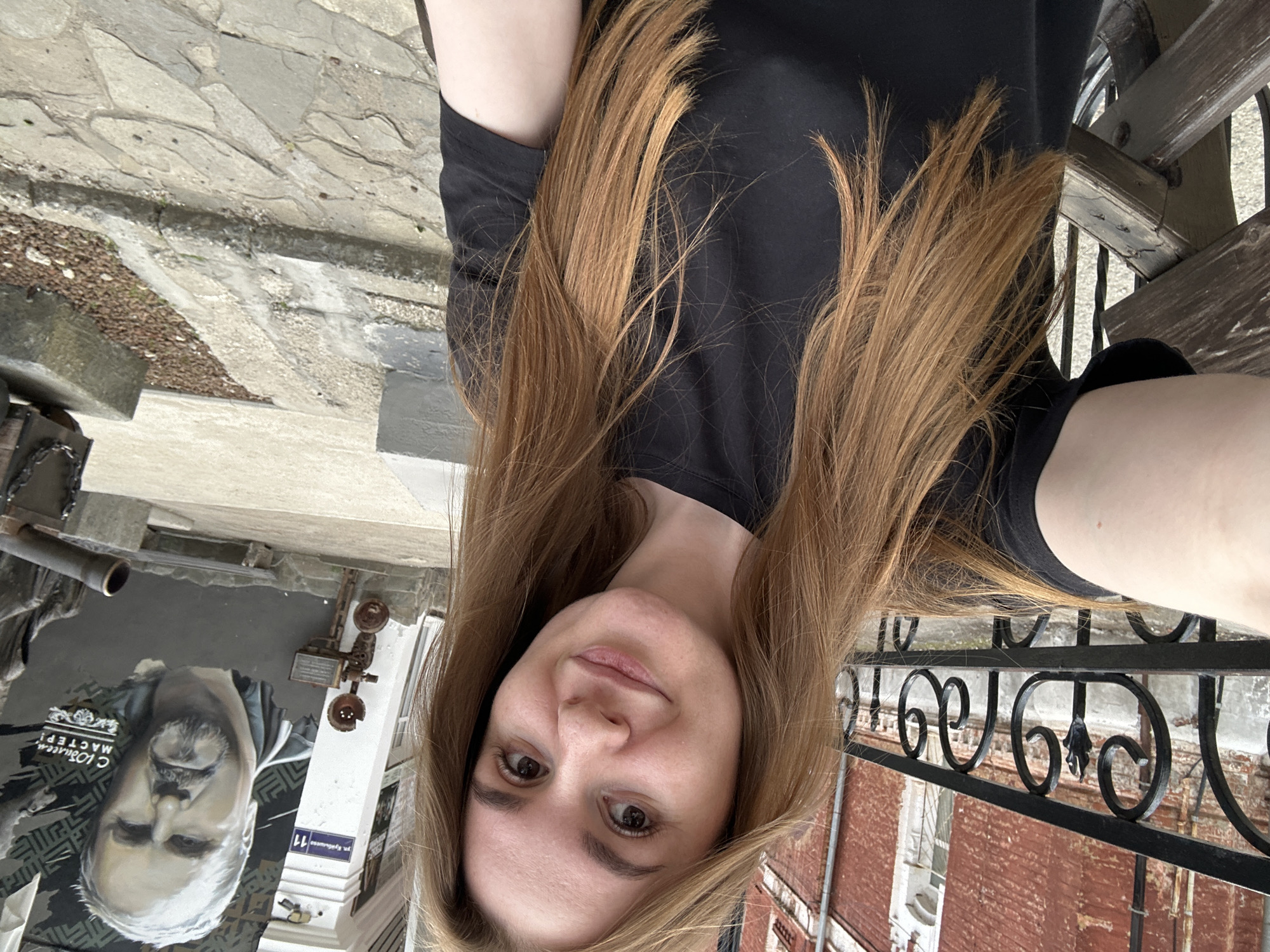 A young woman with long, auburn hair, wearing a black t-shirt, takes a selfie outdoors. She is smiling gently at the camera, her head slightly tilted. The background suggests an urban environment with a brick building on the left, a metal railing, and a mural on a building to the right. The mural features a portrait of a man with a beard and text in Russian that reads "С Юбилеем, МАСТЕР!" (Happy Anniversary, Master!). A street sign in Russian, "ул. Куйбышева 11" (Kuibysheva St. 11), is visible on the building with the mural. The ground is paved with stone and gravel. In the foreground, a wooden bench is partially visible. The overall scene appears to be in Perm, Russia, based on the street name on the mural.