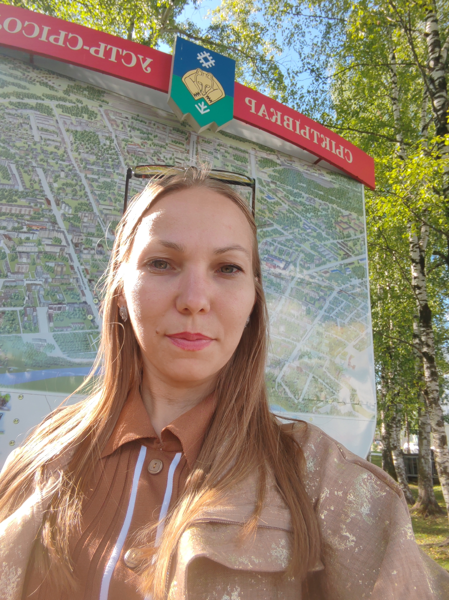 A woman with shoulder-length blonde hair takes a selfie in front of a large map outdoors. The map appears to be a city map, possibly of Syktyvkar, Russia, given the visible text. She is wearing a brown collared shirt with white stripes and a tan jacket with a shimmering, textured pattern. She has on silver earrings and her makeup includes pink lipstick. Behind her, a red sign with Cyrillic text is visible, along with a coat of arms featuring a bear. The background includes trees with green foliage and a blue sky. Sunlight is casting shadows and highlights on her face and clothing.