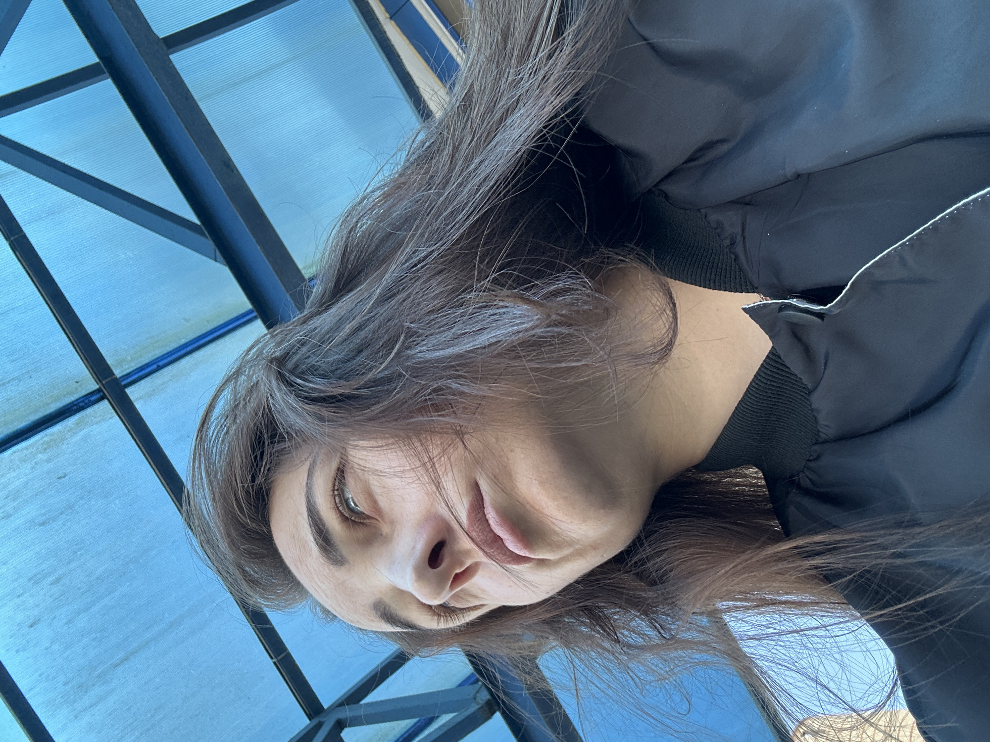 A low-angle, medium shot shows a woman's face and upper body from the shoulders up. She has long, dark brown hair that is windswept and covers part of her face. Her skin is light-toned with some visible freckles. She is looking up and to her left. Her lips are slightly parted and appear to be painted a light pink color. She is wearing a black bomber jacket with a ribbed collar. The background is a bright blue, translucent canopy supported by dark metal beams. The scene appears to be outdoors, possibly under a covered walkway or a bus stop. The image is a selfie, taken from a perspective looking up at the woman. The location is Chekhov, Russia.
