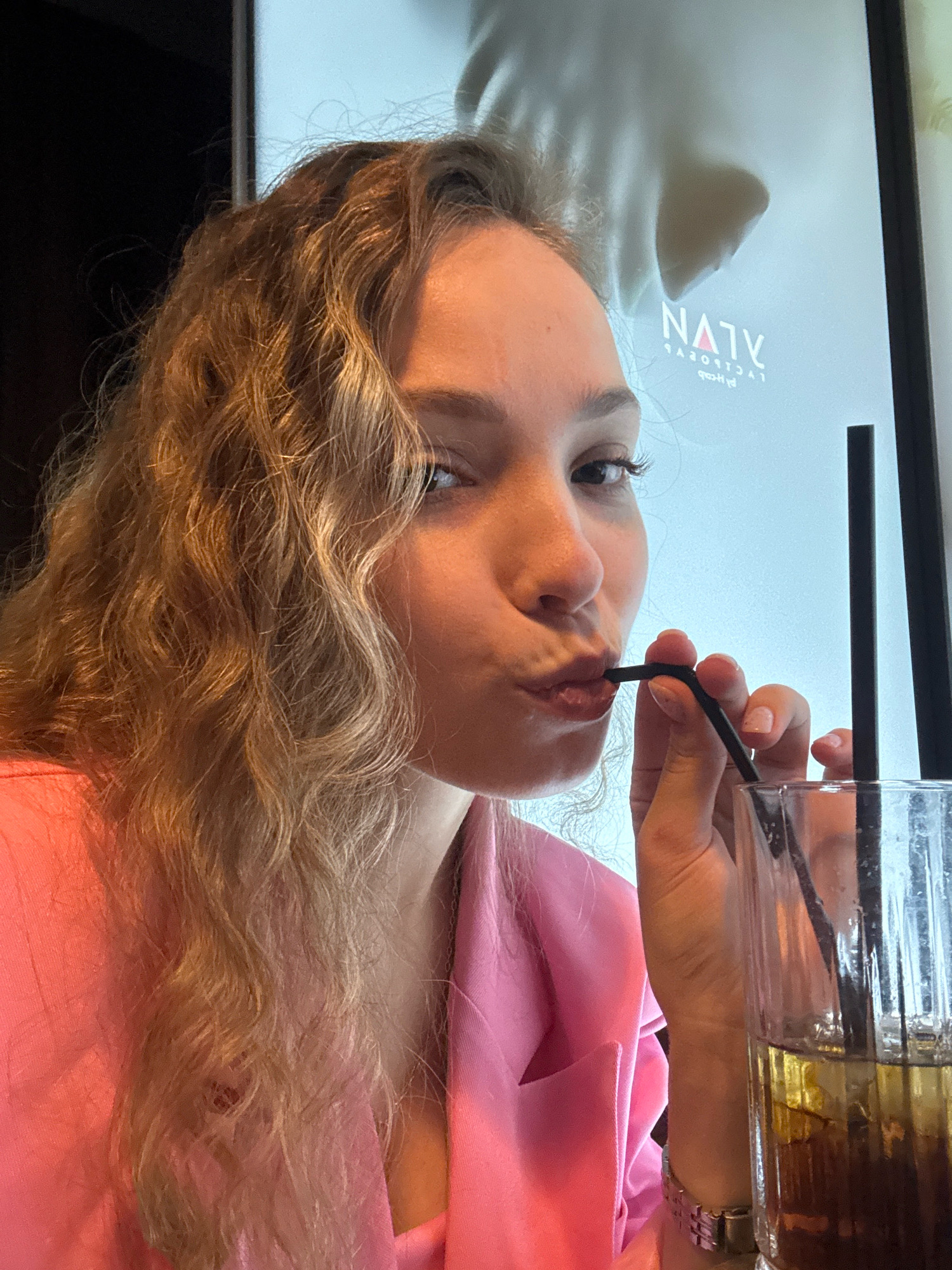 The image shows a young woman with blonde, curly hair wearing a pink blazer, drinking from a glass with a straw. She is looking at the camera with her lips pursed. Behind her, a large poster with text is visible. The scene appears to be indoors, possibly in a cafe or restaurant. The woman is holding the straw with her right hand, and her left hand is resting on the rim of the glass. The glass contains a dark liquid with ice.