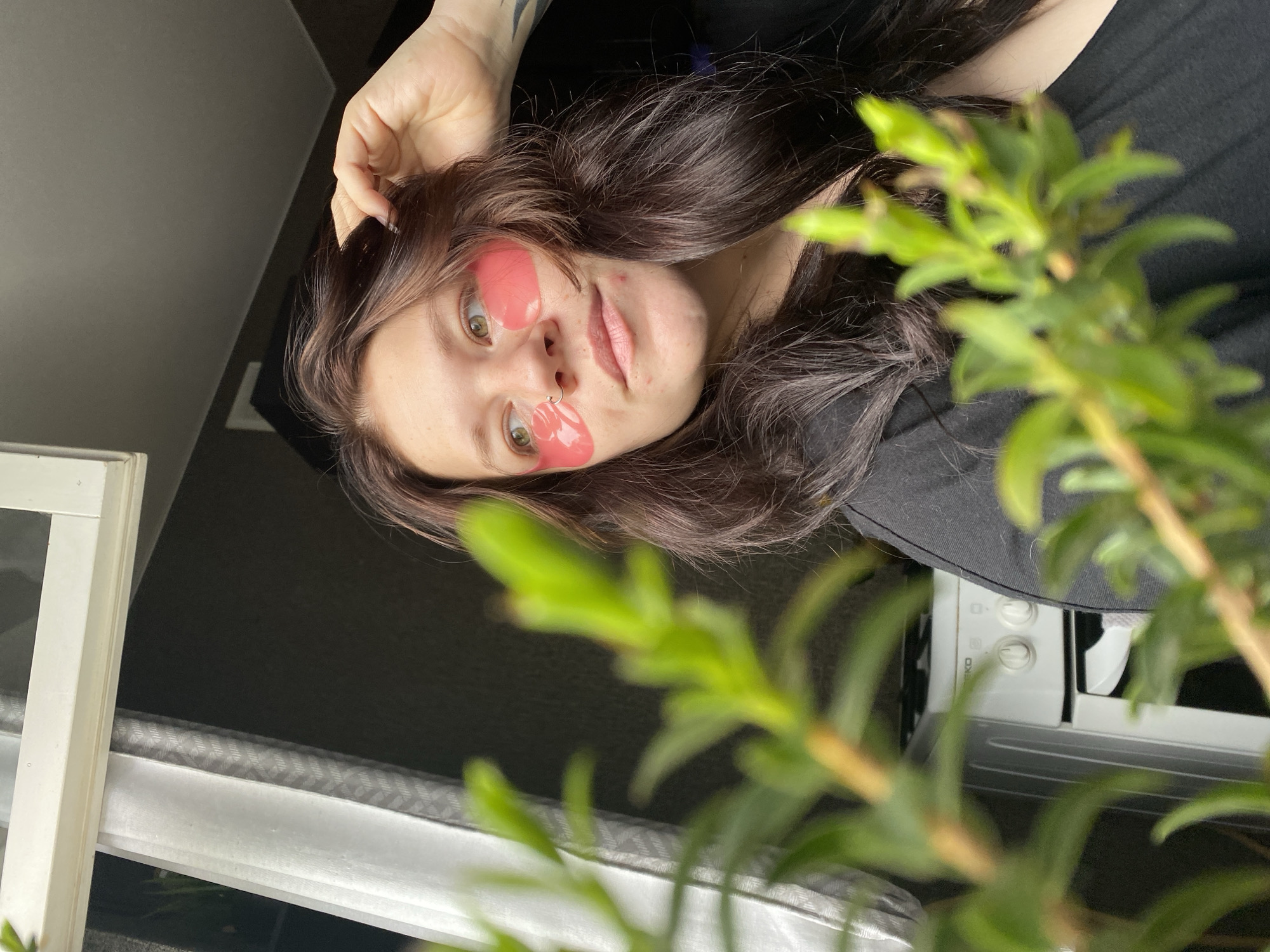 This image, taken in Chaikovsky, Russia, features a young woman lying down and looking upwards towards the camera. She has long, dark brown hair that frames her face. Her eyes are light-colored, possibly green or hazel, and she has a small silver ring piercing in her left nostril. Pink, tear-shaped hydrogel patches are applied to her face, specifically on her upper cheekbones and beneath her eyes. Her expression is neutral to slightly pensive, with her lips slightly parted. Her skin shows some minor blemishes on her forehead and cheeks. One of her hands is visible near her head, lightly touching her hair, and a tattoo is faintly visible on her arm. She is wearing a dark, likely black, top.
The foreground of the image is dominated by out-of-focus green leaves and stems of a plant, suggesting it is positioned between the camera and the subject. Through the blurred plant on the right side of the frame, a white appliance with two circular knobs and the partially visible text "SEKO" can be seen, possibly a microwave or an oven. To the left, a white, vertical structure, resembling a window frame or part of a furniture piece, is visible. The background behind the woman is dark and indistinct, creating depth. The lighting appears to be natural, indicating it is daytime. The overall scene suggests a relaxed, casual moment of self-care in an indoor setting.