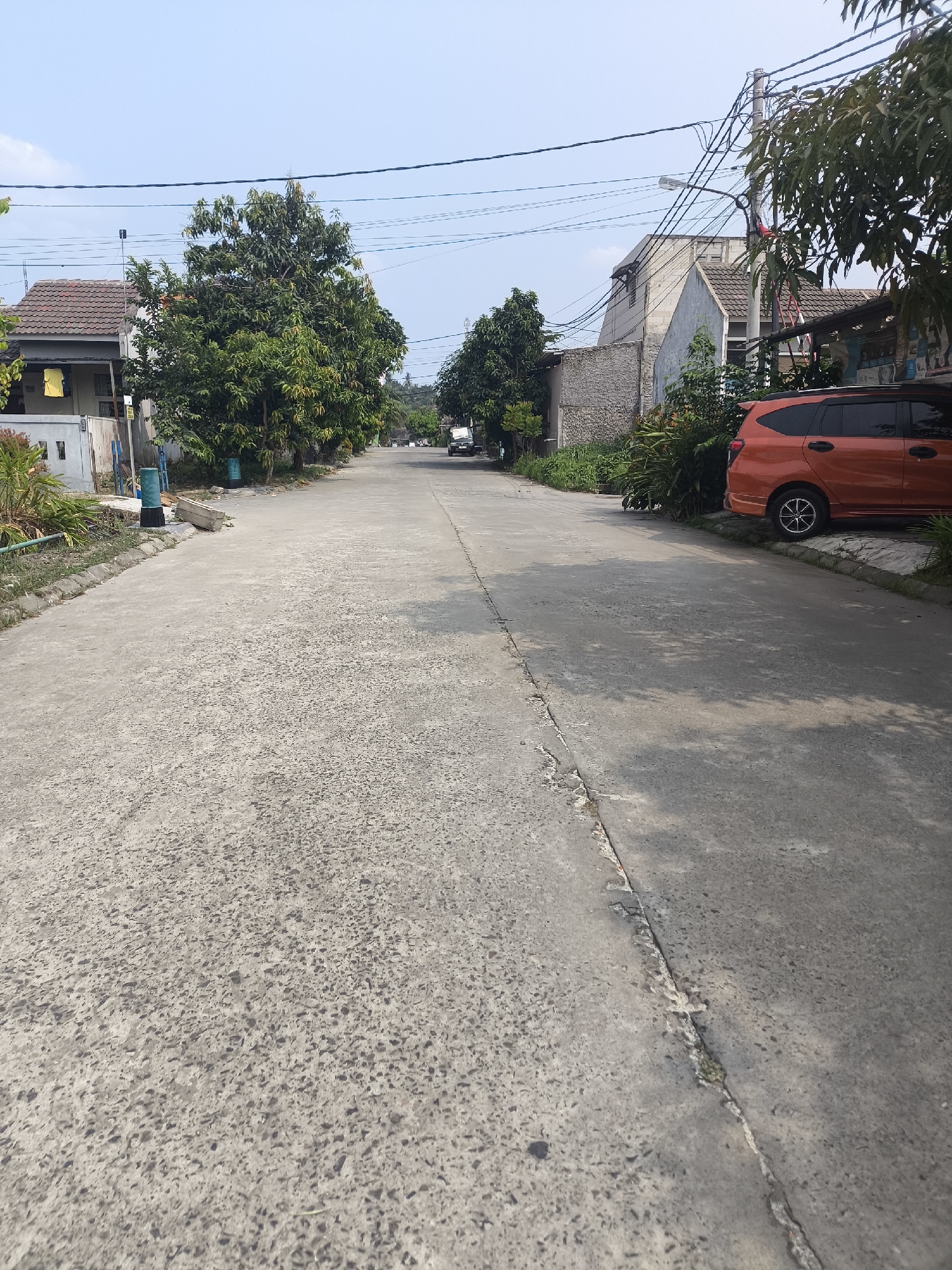 The image depicts a street in a residential area of Cilegon, Indonesia, under a clear, bright sky. The road itself is paved with concrete, showing signs of wear and tear with visible cracks and uneven textures. On the left side of the street, a house with a brown tiled roof is partially visible, along with some greenery and a yellow cloth hanging outside. Further down the road, several trees line the left side, their leaves casting dappled shadows on the pavement.
On the right side of the street, an orange minivan is parked. Behind it, there is dense foliage and more trees. The background reveals more houses and buildings, some appearing to be under construction with exposed brickwork. Overhead, a network of power lines and utility poles crisscross the sky, a common sight in urban and suburban environments. The overall impression is a typical residential street scene in a tropical climate, with the characteristic architecture and vegetation of Indonesia.