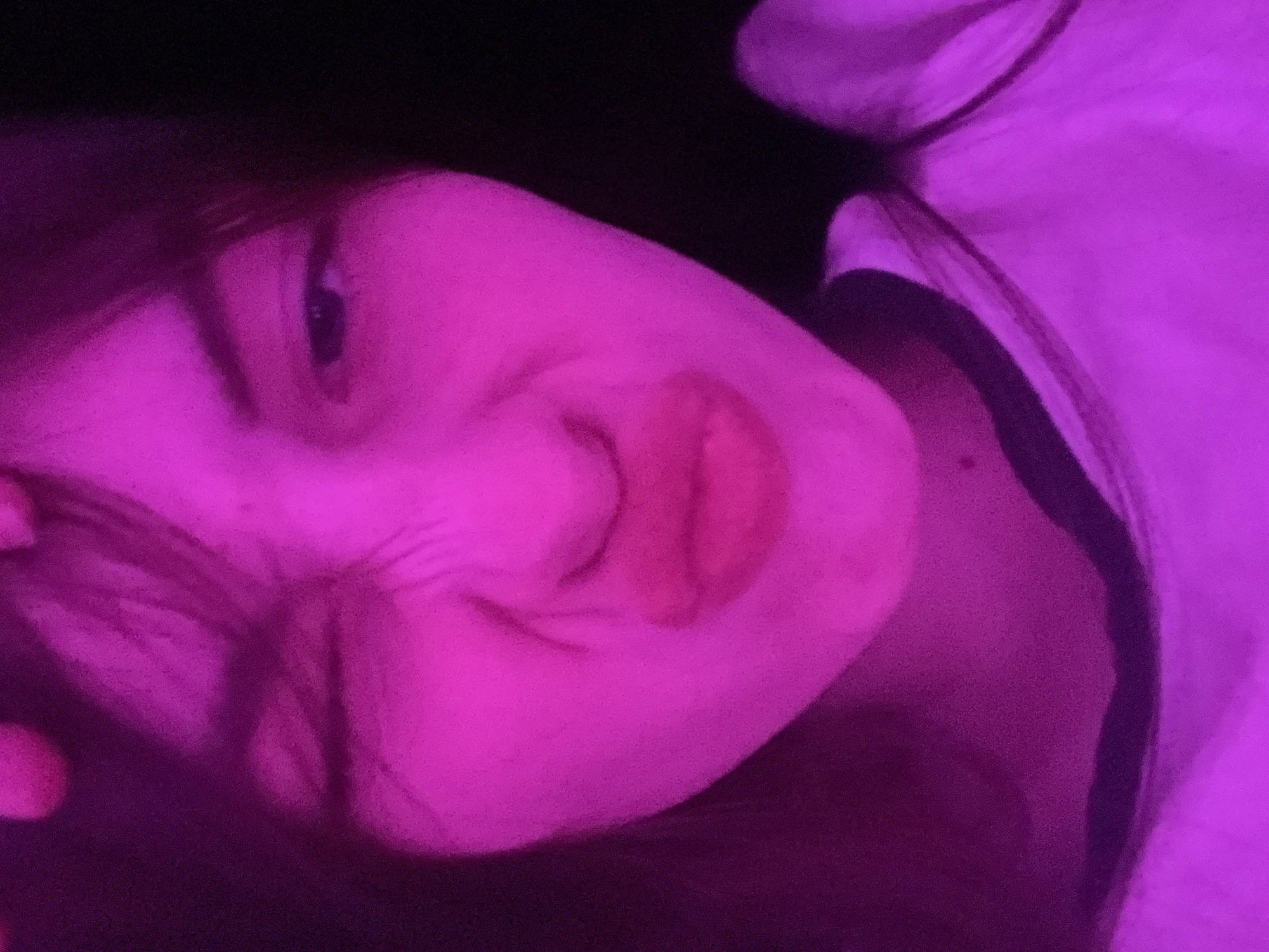 The media file features a close-up portrait of a person, likely a young woman, lying down in a dimly lit indoor setting. The scene is bathed in a strong, vibrant magenta or purple light, casting deep shadows and highlighting the person's features.
The subject's face is central to the image, viewed from an upward angle, with their head tilted. One eye is visible, looking towards the camera. A subtle smile or smirk plays on their lips. A distinctive detail is the red or pink lipstick, which is noticeably smeared around the mouth and onto the cheek, giving a playful or disheveled appearance. Dark hair frames the face, blending into the dark background. On the right side of the frame, a portion of white fabric, possibly clothing or bedding, is visible.
The activity appears to be the person relaxing or posing for a picture in a low-light environment. The strong colored lighting suggests a night-time setting or an intentional artistic lighting setup, possibly from a neon light or LED. No specific location cues point to Saint Petersburg, Russia, beyond the provided context. There is no visible text in the image.