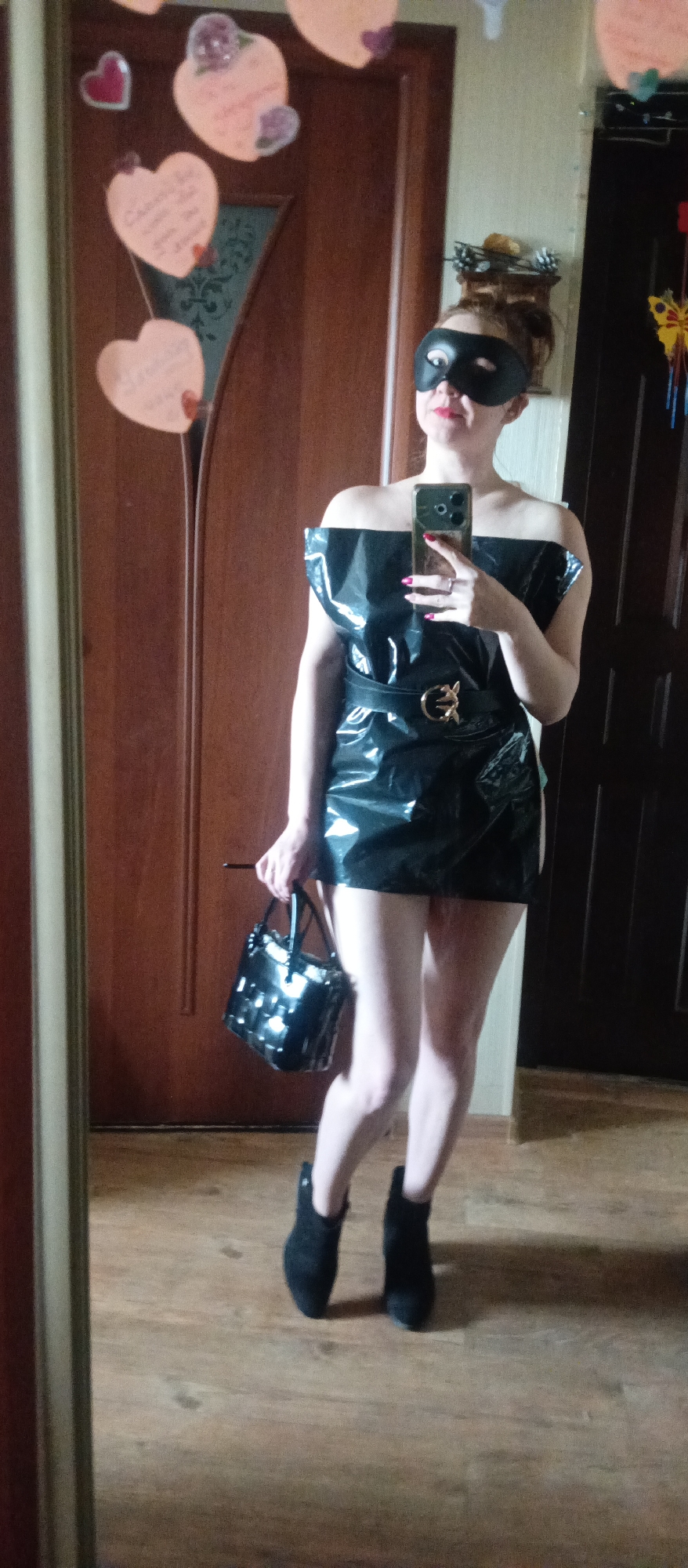 A woman is captured in a mirror selfie, standing indoors and posing. She wears a black, off-shoulder dress with a shiny, possibly latex or PVC, finish. A black belt with a prominent gold-colored buckle cinches her waist. Her legs are bare, and she wears black ankle boots. Her face is partially obscured by a black eye mask, but red lipstick is visible on her lips, suggesting a slight smile. She holds a black, shiny handbag in her left hand and a smartphone, likely for taking the picture, in her right hand.
The background shows a warm-toned wooden door with a decorative glass panel. Several heart-shaped decorations, some with handwritten text in light colors, are suspended on or near the door. The text is largely illegible, but some cursive writing is visible. To the left, a portion of the mirror frame is visible. The floor is made of wooden planks. The lighting suggests an indoor setting, possibly a home.