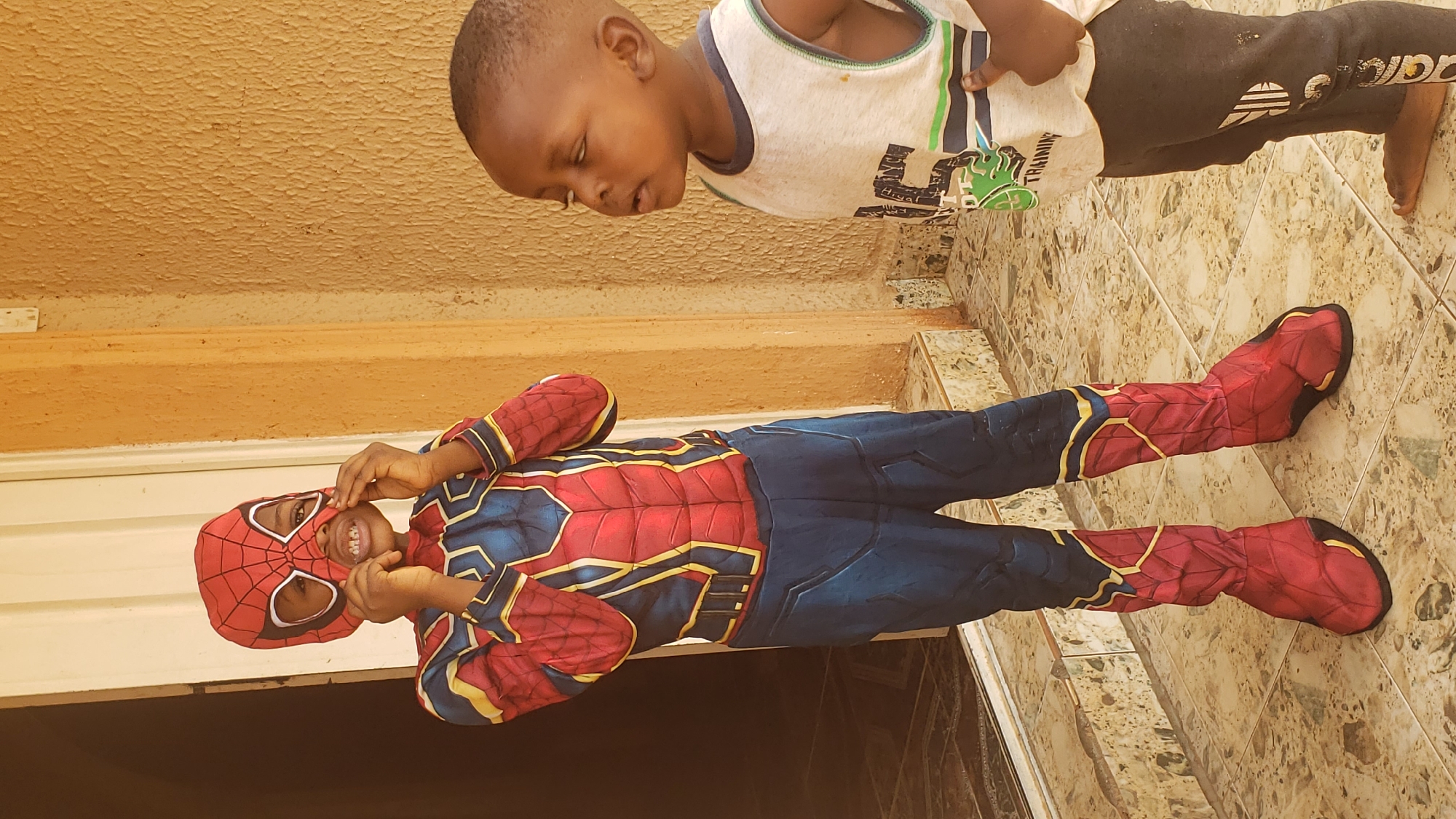 Two children are positioned in an outdoor setting. One child is dressed in a Spider-Man costume, complete with a mask, suit, and attached boots. The costume is red and blue with gold accents, and the child is smiling, pulling the mask slightly away from the face. The second child, not in costume, is wearing a tank top and dark sweatpants and is positioned above the Spider-Man figure, looking down. The background consists of a tan-colored wall, a white trim, and a tiled floor. The location is Sabon Tasha, Nigeria.