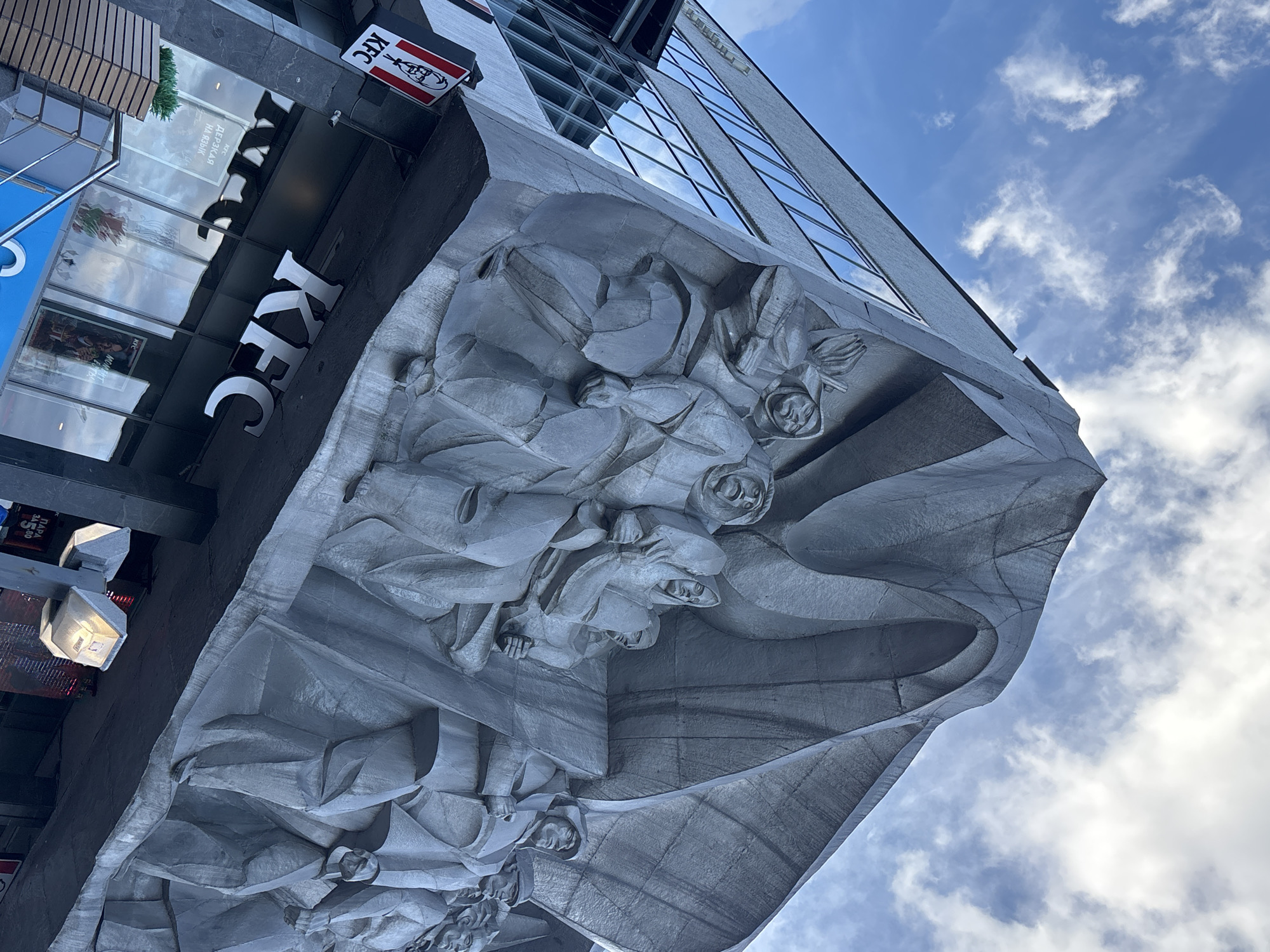 The image depicts the facade of a building in Minsk, Belarus, featuring a large, monumental bas-relief sculpture. The sculpture, rendered in gray concrete or stone, depicts a group of figures in a dynamic, marching pose, possibly representing a historical or ideological theme. The figures are stylized and appear to be wearing period clothing, including hats and uniforms, with some figures carrying what may be flags or banners.
Below the sculpture, the building's lower levels house a KFC restaurant. The entrance to the KFC is visible, with its distinctive red and white logo prominently displayed. Large glass windows reflect the sky and the surrounding environment. The sky above is blue with scattered white clouds, suggesting daytime. The architectural style of the building appears to be from the Soviet era, common in Minsk. The perspective of the photograph is from a low angle, looking upwards towards the sculpture and the sky, emphasizing the scale and grandeur of the artwork. The scene is devoid of any discernible human interaction beyond what is depicted in the sculpture.