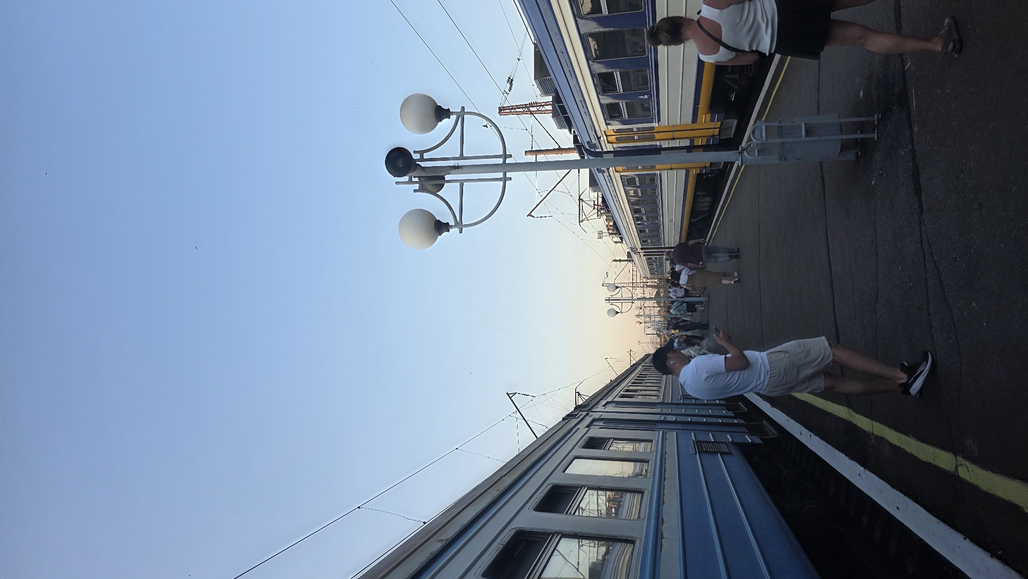 The image depicts a train station platform in Koziatyn, Ukraine, with several trains present. The sky is clear and blue, indicating daytime. Two trains are visible on parallel tracks, one on the left with a blue and light blue exterior, and another on the right with a yellow and blue exterior. A group of people are standing and walking on the platform between the trains. In the foreground on the left, a man wearing a white t-shirt and beige shorts is looking at his phone. On the right, a woman wearing a white tank top and a dark skirt is also looking at her phone. There are several lampposts with multiple lights spaced along the platform. The ground of the platform appears to be asphalt. The overall scene suggests a typical day at a train station.