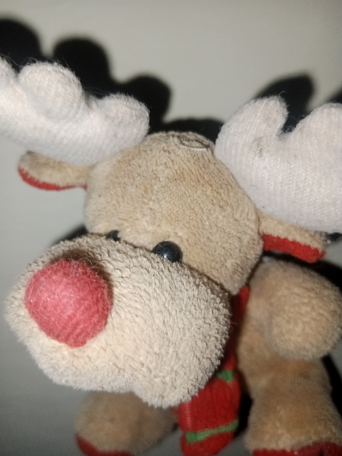 A close-up photograph features a stuffed reindeer toy. The reindeer has a beige, furry face with a large, red, round nose. Small, black, beady eyes are visible. White antlers with red trim stick out from the top of its head. A red and green striped scarf wraps around its neck. The background is a blurry white with dark, indistinct shadows. The overall impression is of a festive and well-loved toy.