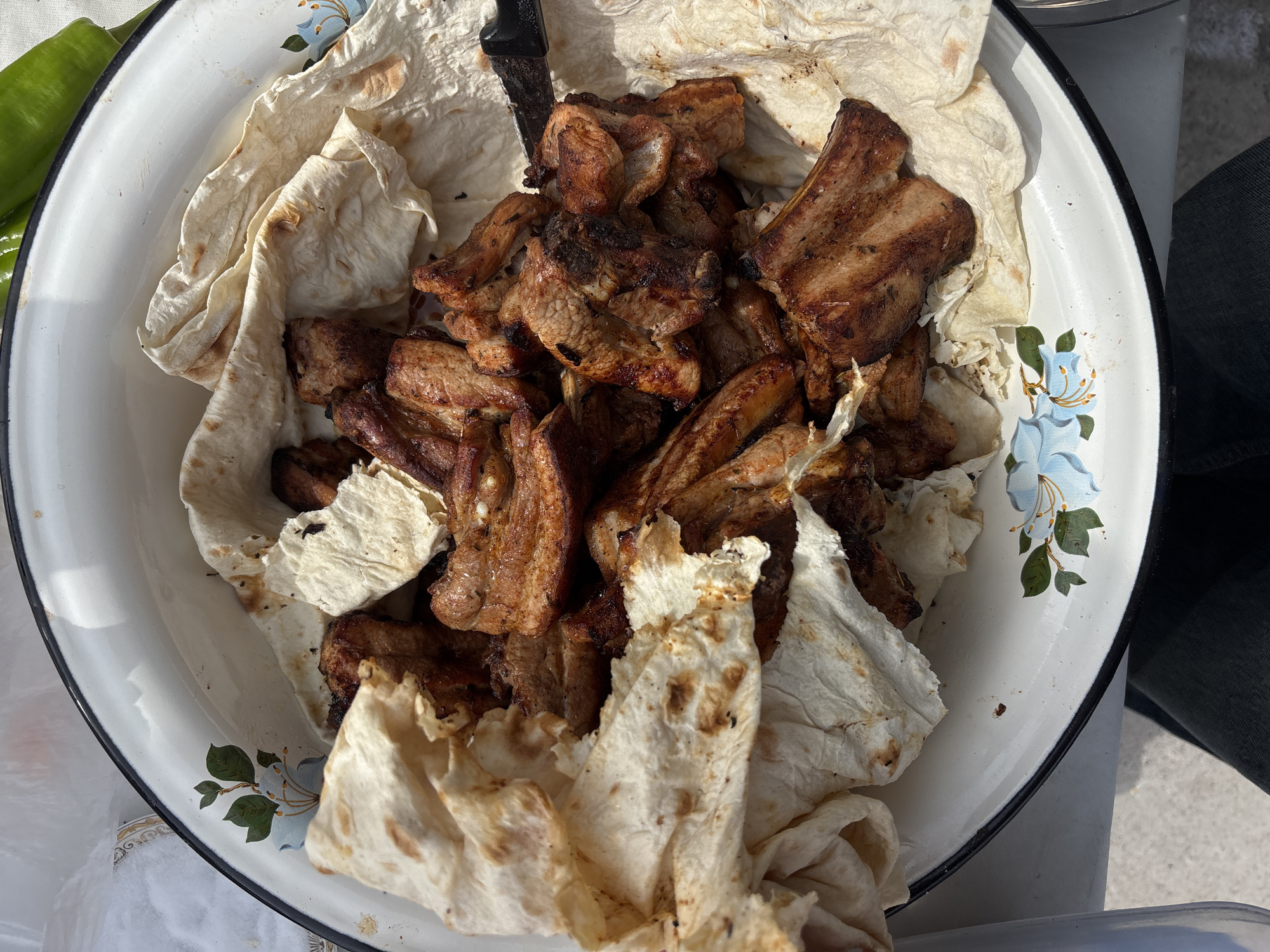 A top-down, eye-level shot shows a white enamel bowl with floral decorations containing grilled meat and lavash (thin flatbread). A black-handled knife is partially visible amidst the food. A green pepper is on the left side of the bowl. The bowl sits on a light-colored surface. A person's leg, clad in dark fabric, is visible in the background on the right. The location is identified as Abovyan, Armenia.