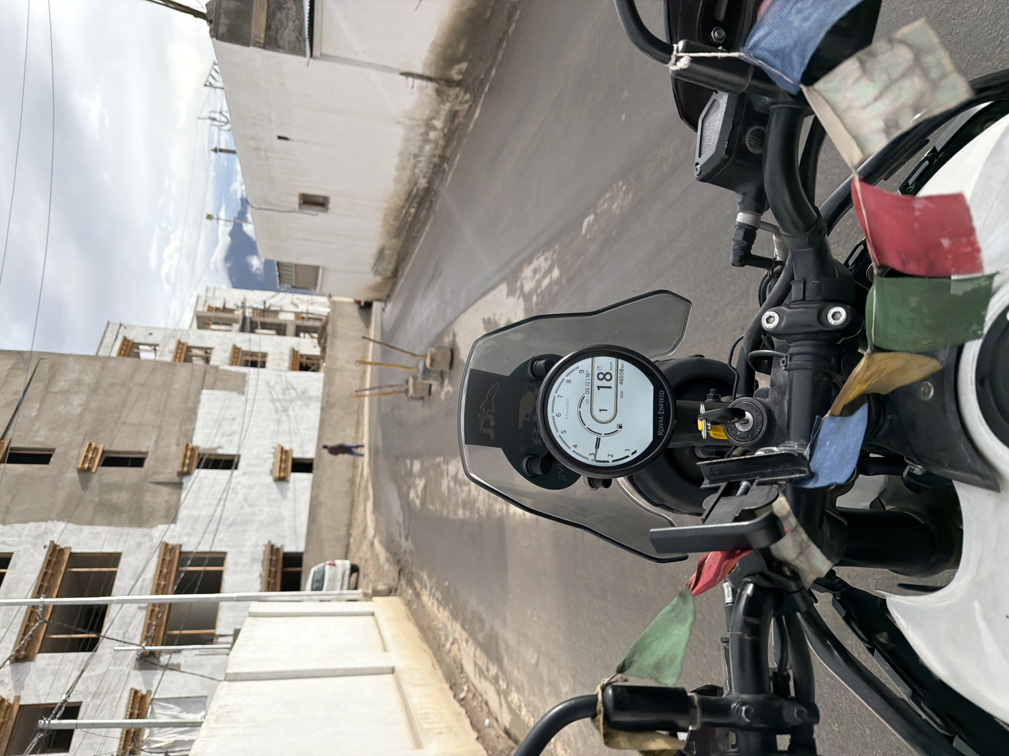 The image depicts a point-of-view shot from a motorcycle in the city of Leh, India. The motorcycle's handlebars, dashboard with a digital display showing "18" and "46508 km", and prayer flags are in the foreground. The road ahead is paved, with a white wall on the left and buildings on both sides. The buildings appear to be under construction or recently built, with white facades and wooden window frames. In the distance, mountains are visible under a cloudy sky. A white car and a person walking are present on the left side of the road. The overall scene suggests a street in a mountainous region, consistent with Leh.